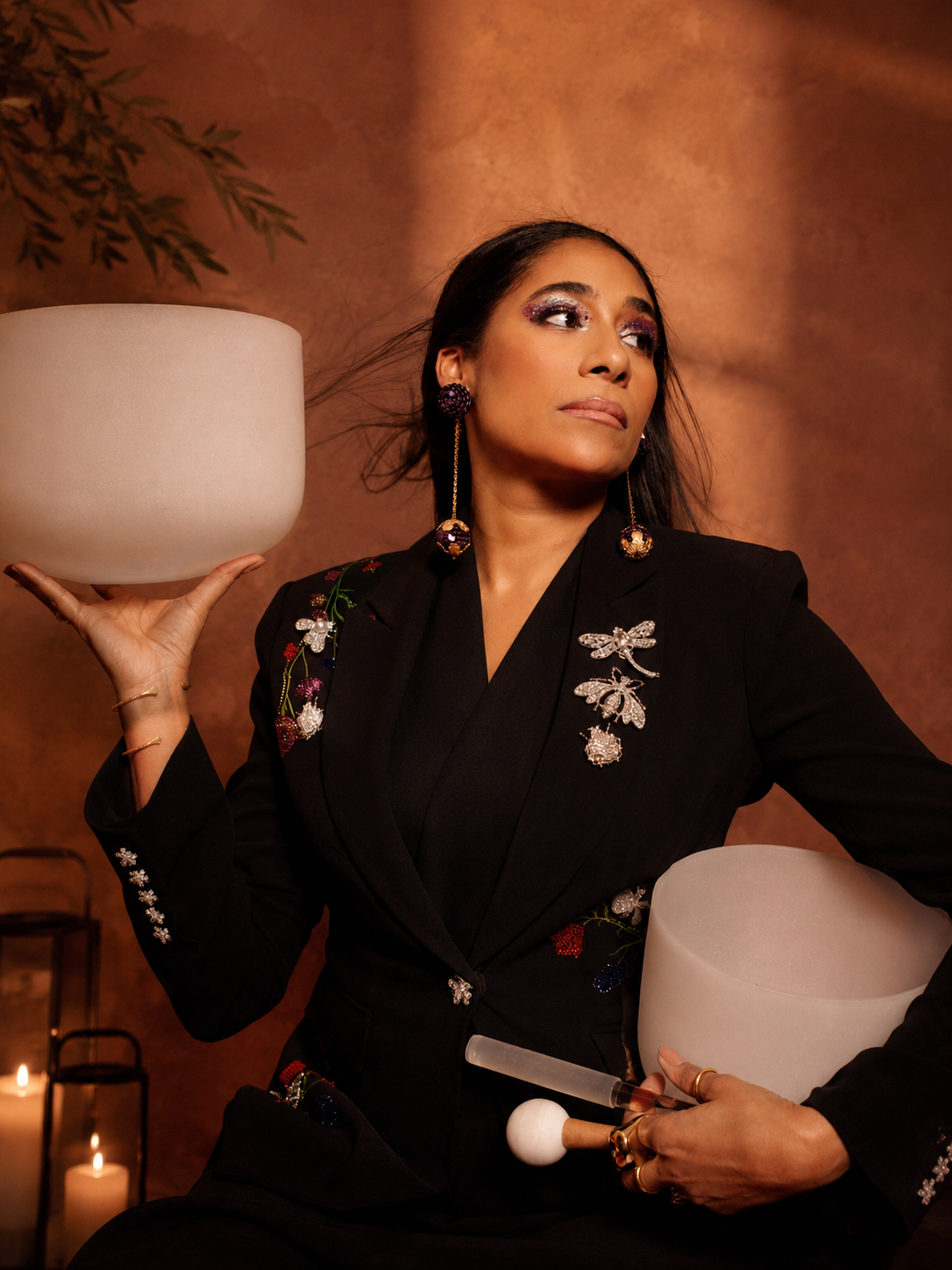 A woman with long dark hair, wearing a black blazer with embroidered and jeweled floral designs, holds a white percussion instrument and a mallet, with a serene expression against a warm-toned background.