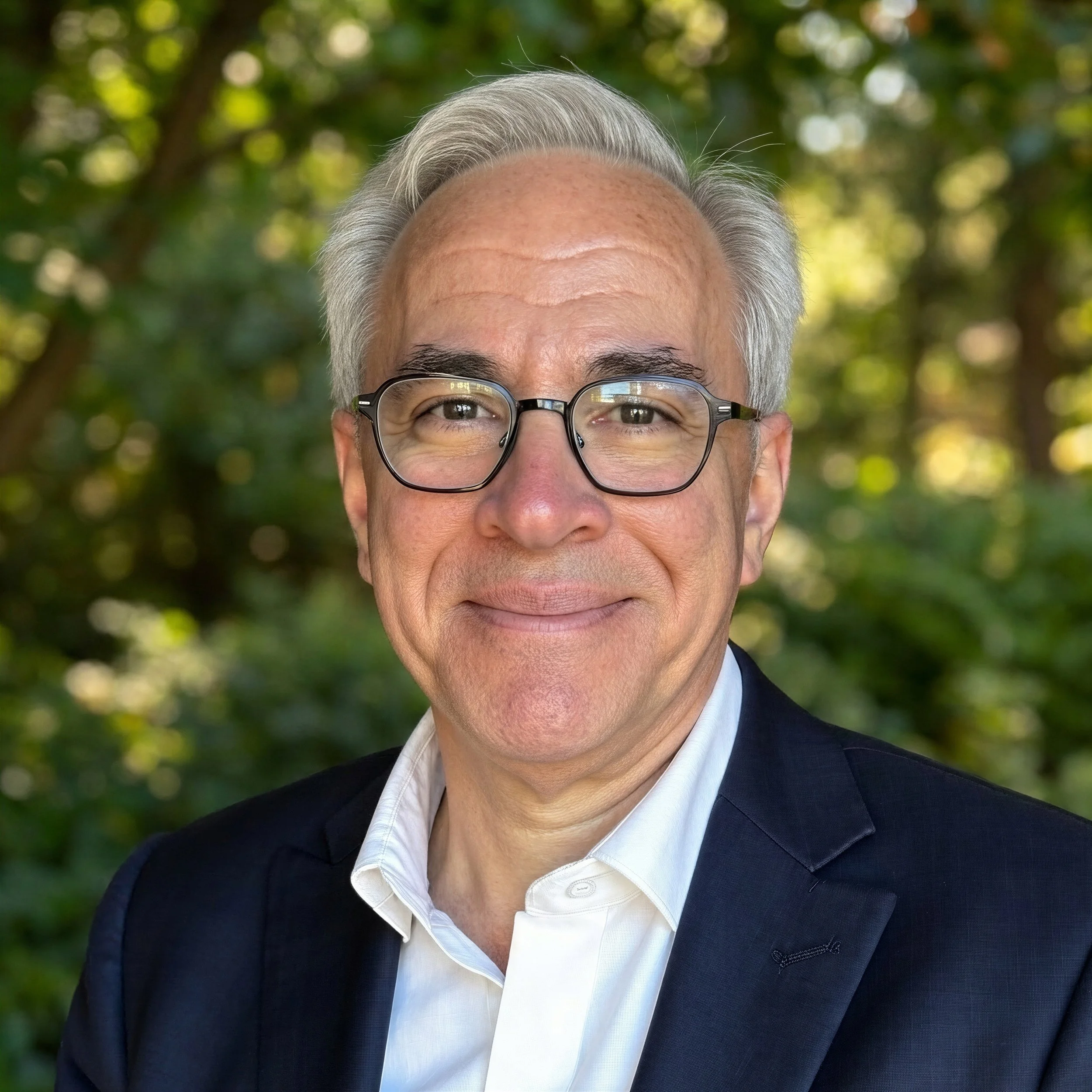 A middle-aged man with gray hair, glasses, and light skin, smiling outdoors with a blurred green leafy background, wearing a dark blazer and white shirt.