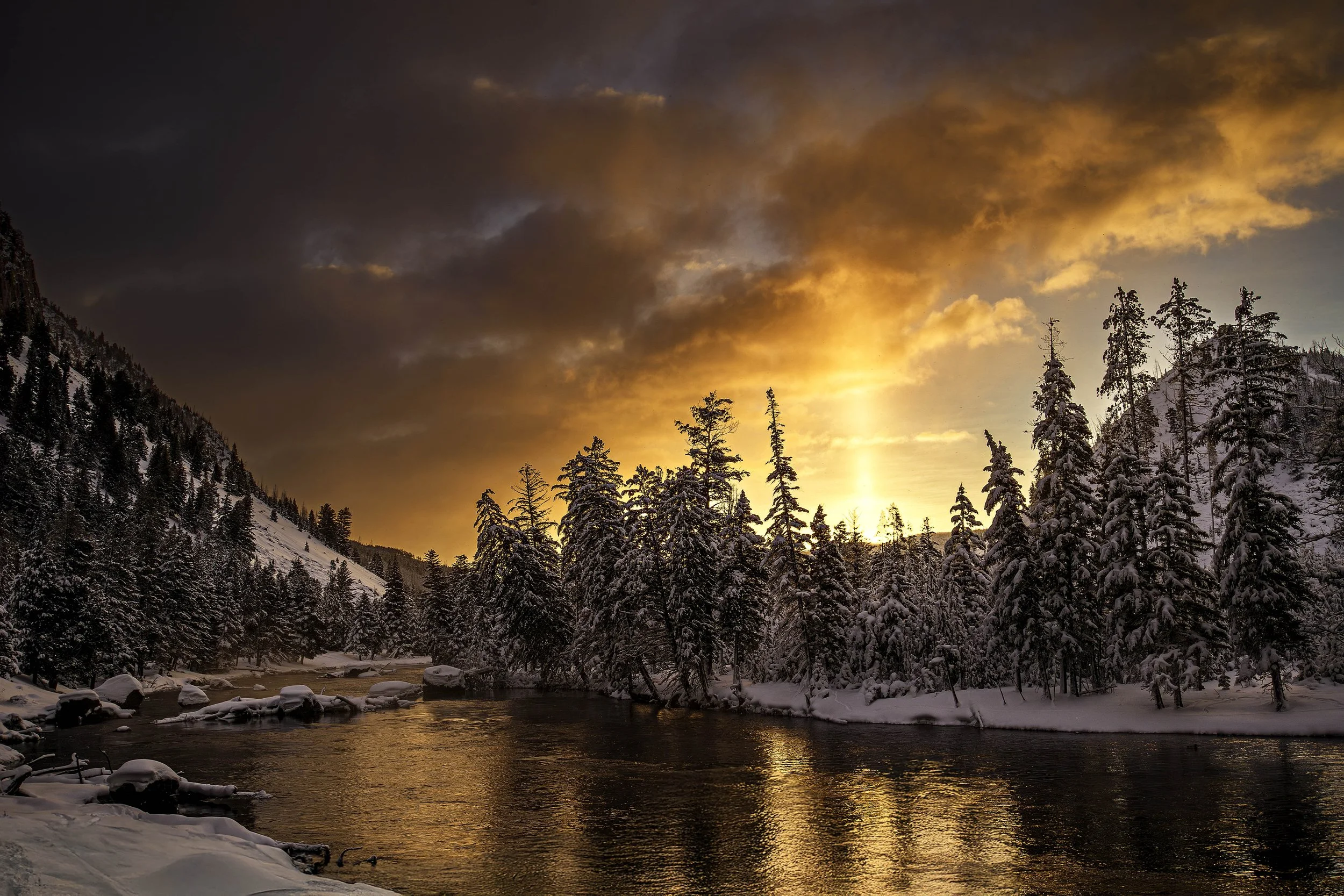 A snowy river landscape at sunset with tall pine trees along the banks and mountains in the background, sky filled with orange and purple clouds.