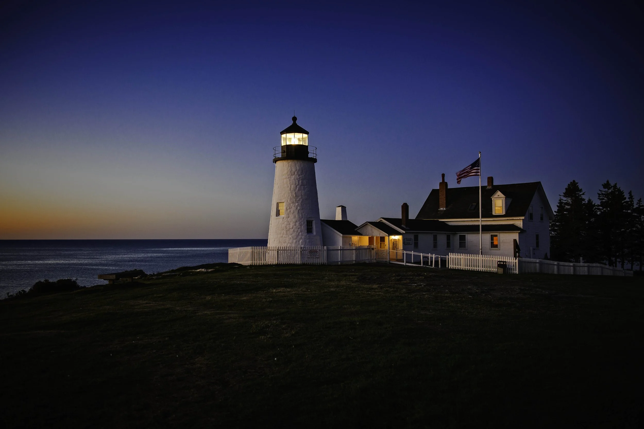 Lighthouse overlooking the ocean at dusk with a house nearby and an American flag on a flagpole.