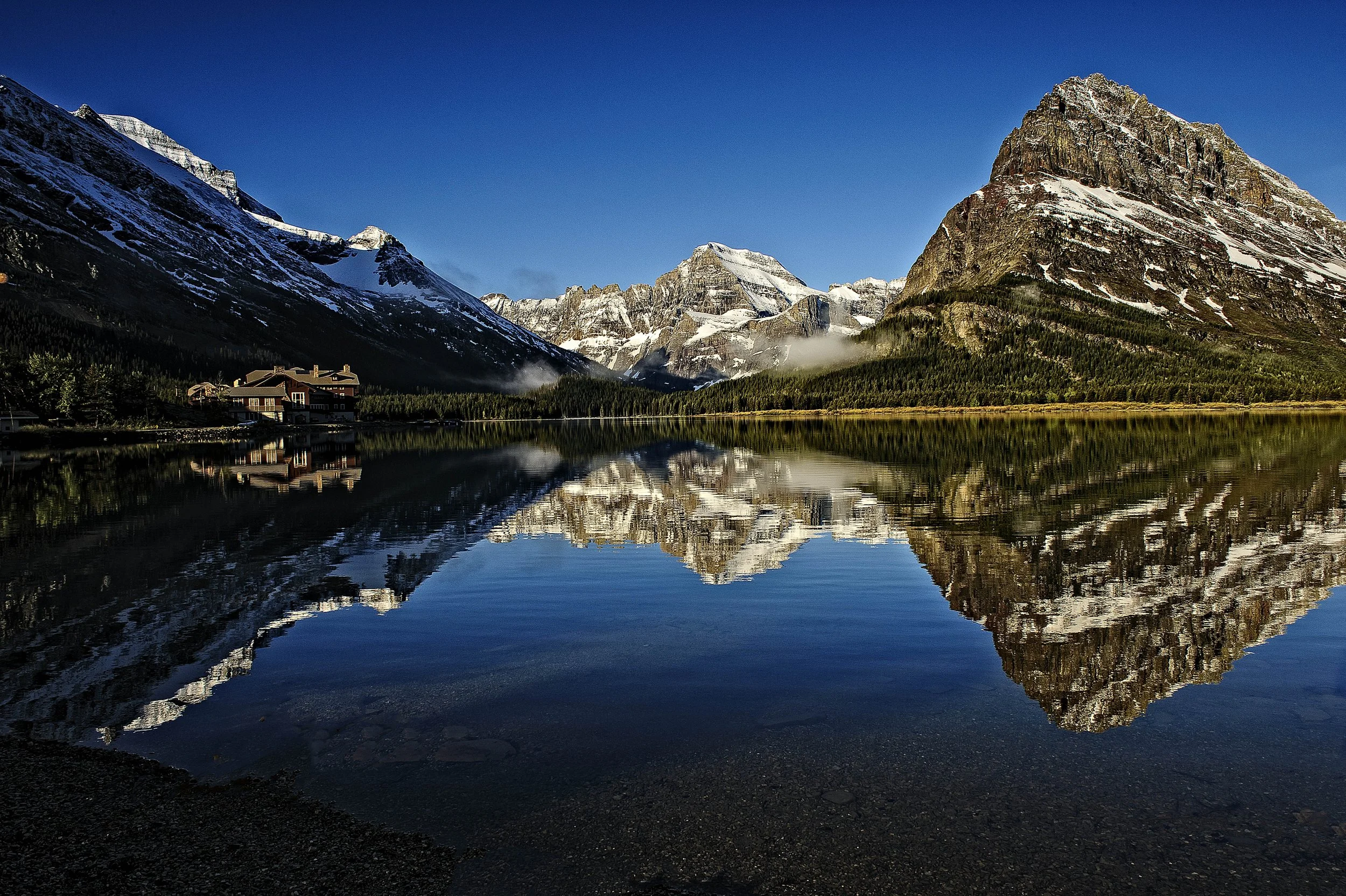 Mountain landscape with snow-capped peaks and a clear lake reflecting the mountains, forest, and a building on the shoreline under a blue sky.