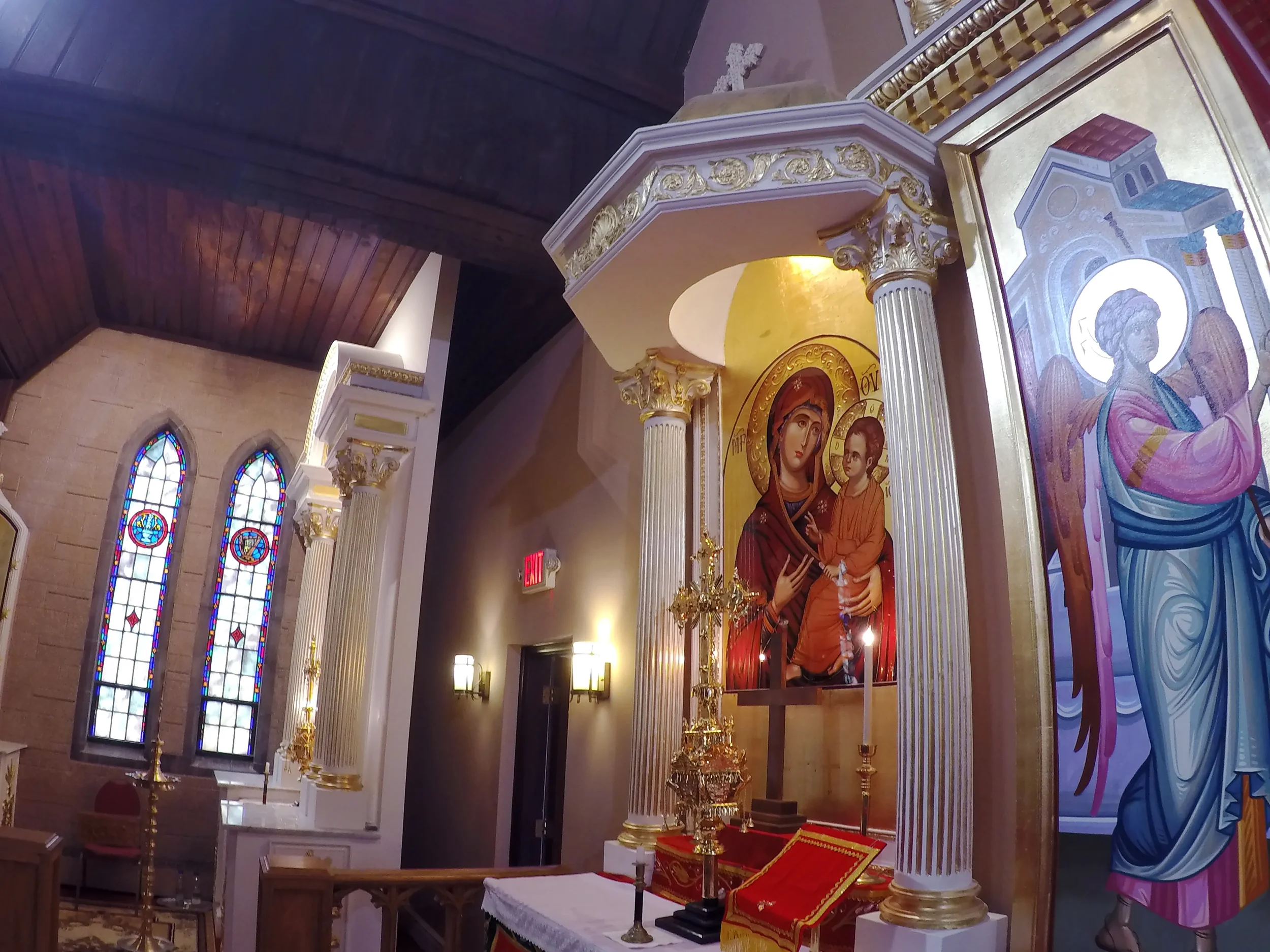 Interior of a church featuring stained glass windows, religious icons, and ornate columns with gold accents. The central icon depicts Mary holding baby Jesus.