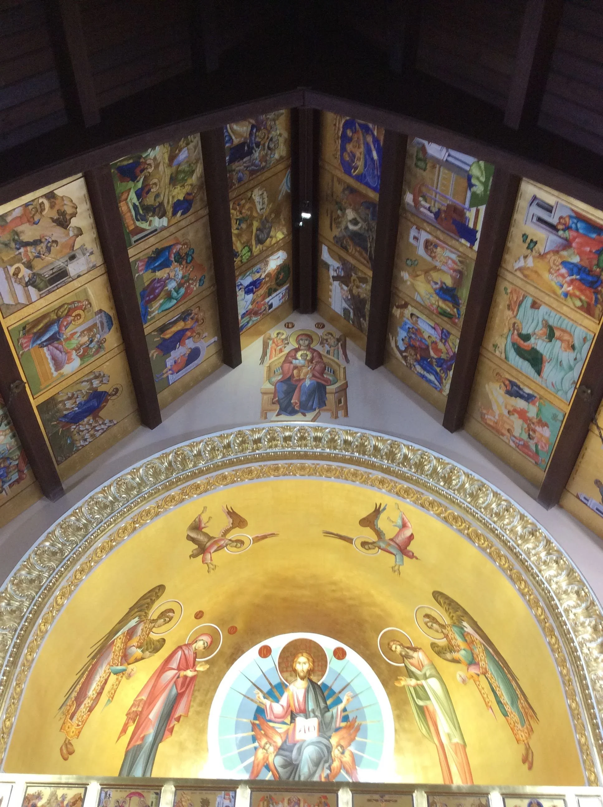 Interior view of a church with a large religious icon mural of Jesus Christ at the altar, surrounded by angels, and ornate gold and colorful paintings of biblical scenes on the walls.