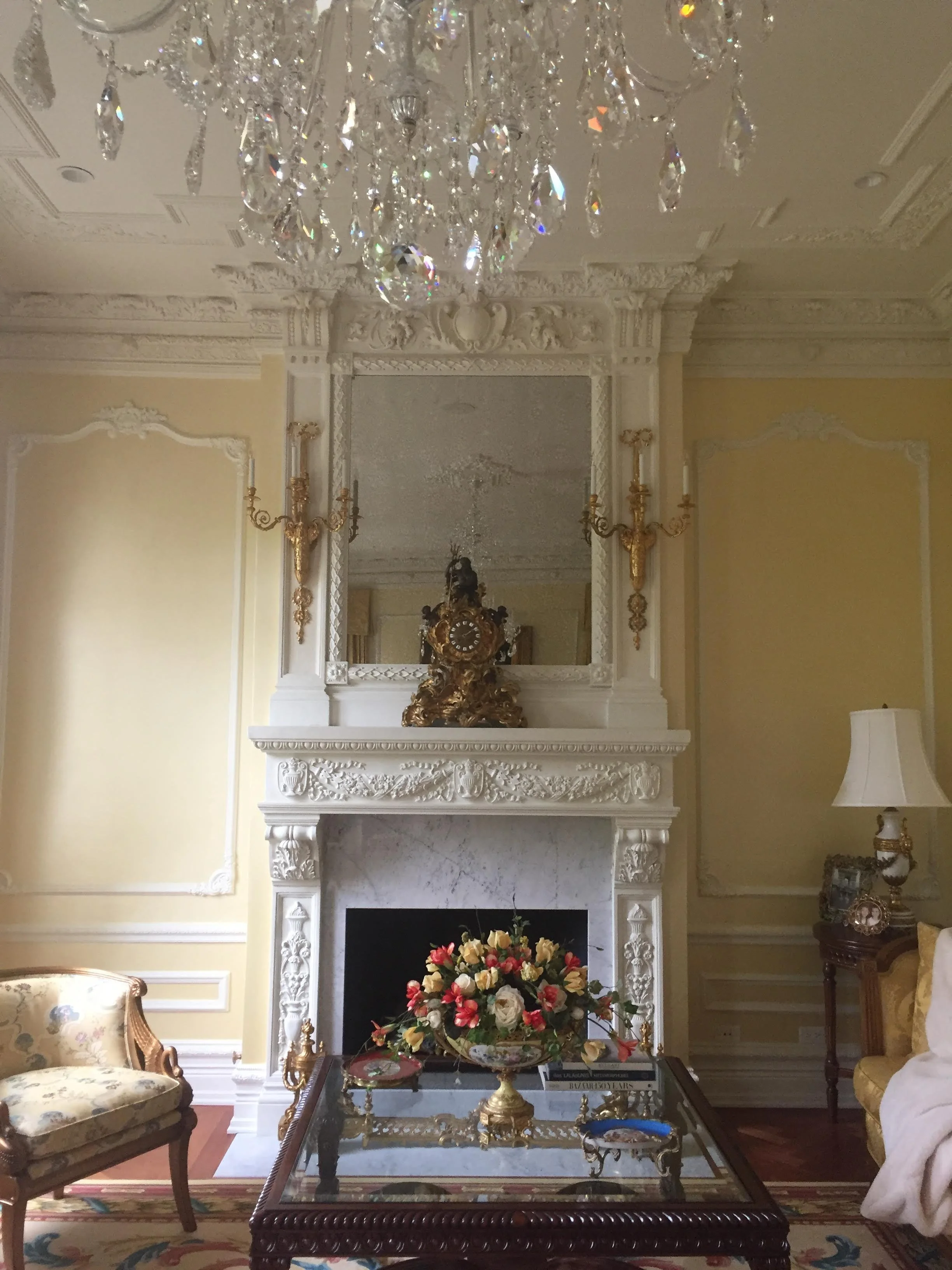 Elegant living room with a white ornate fireplace, a large mirror above it, and decorative gold and white details on the walls. A crystal chandelier hangs from the ceiling. In front of the fireplace, there's a glass-top table with a gold vase filled 
