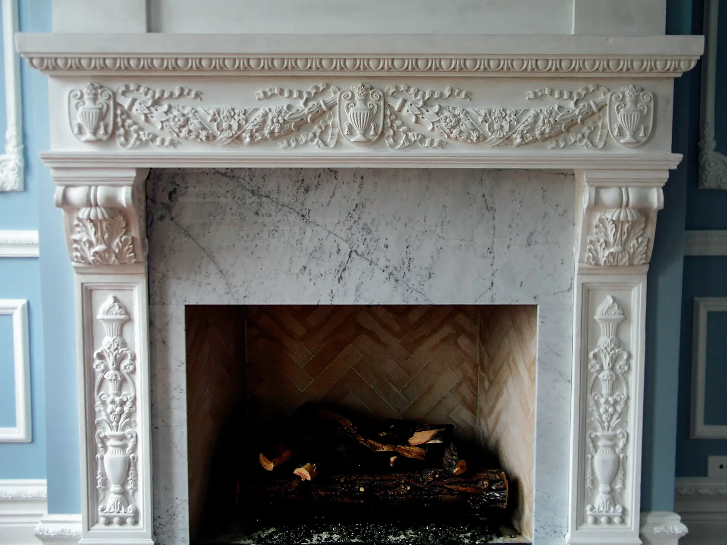 An ornate white marble fireplace with intricate floral and urn decorative carvings on the mantel and sides, with logs inside.