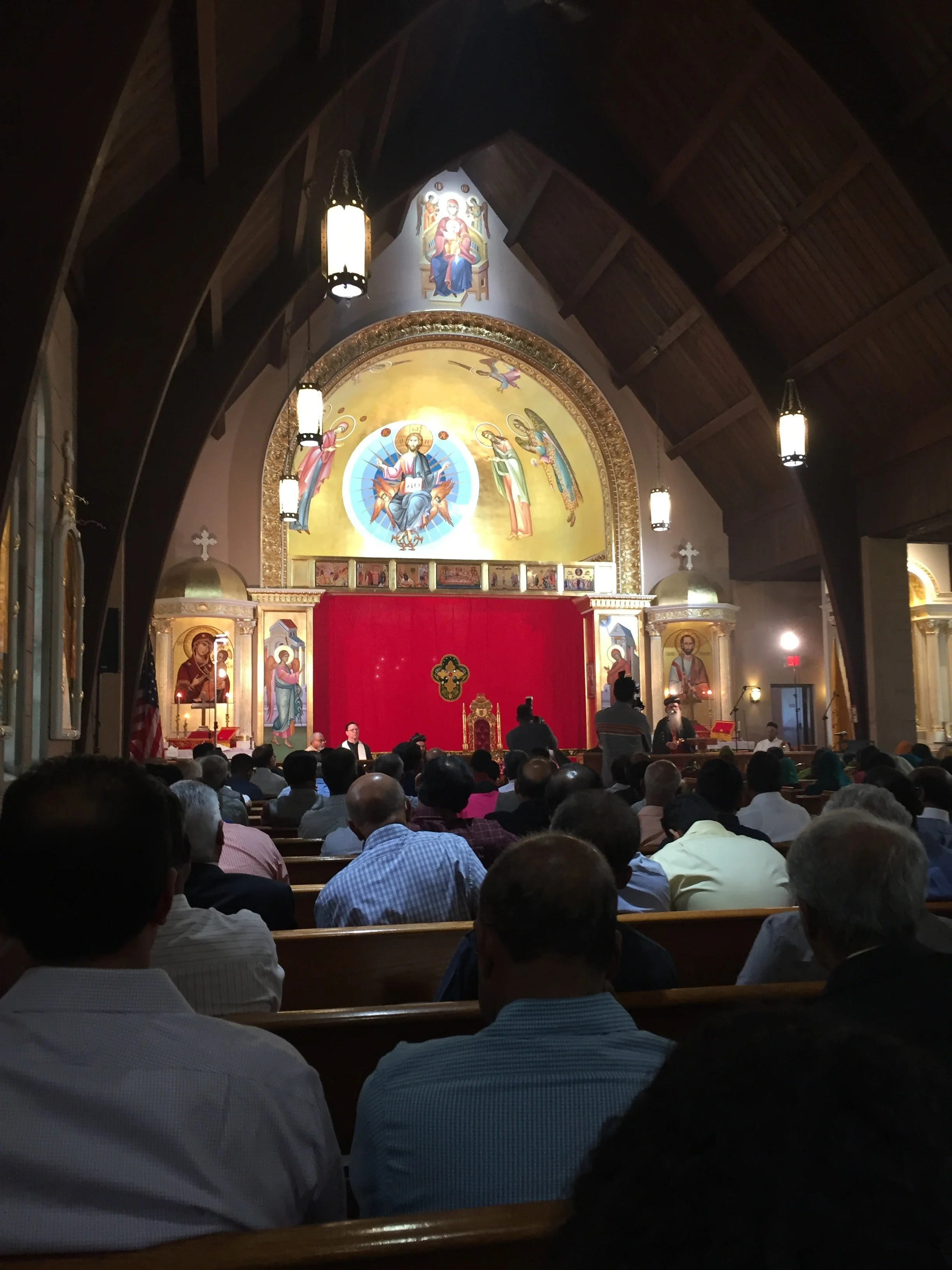 Inside a church with a large congregation, overlooking the altar with religious icons, paintings, and chandeliers hanging from a wooden ceiling.