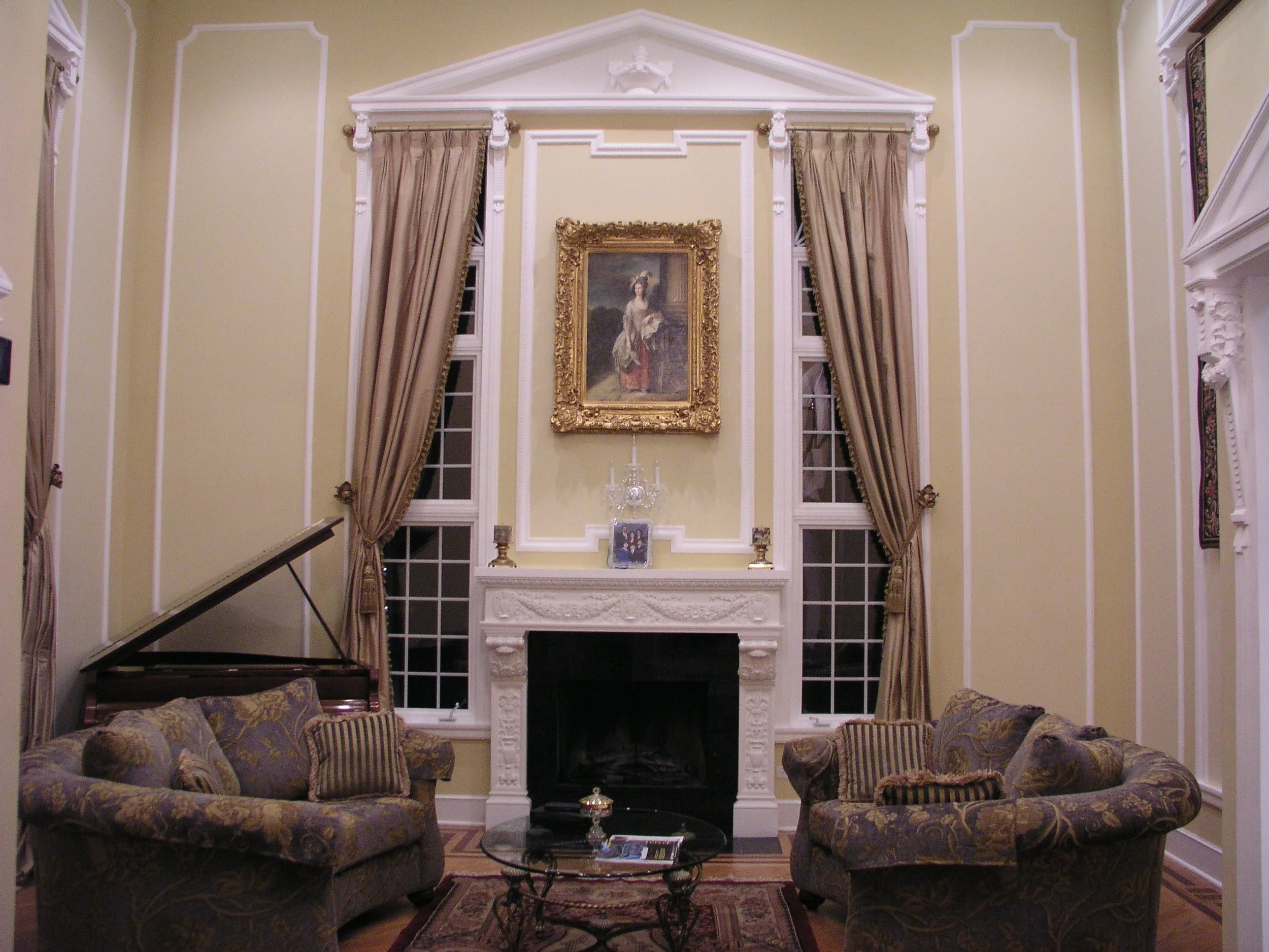 Elegant living room with high ceilings, ornate crown molding, a fireplace, two large windows with gold curtains, a grand portrait above the mantel, and vintage sofas arranged around a glass coffee table.