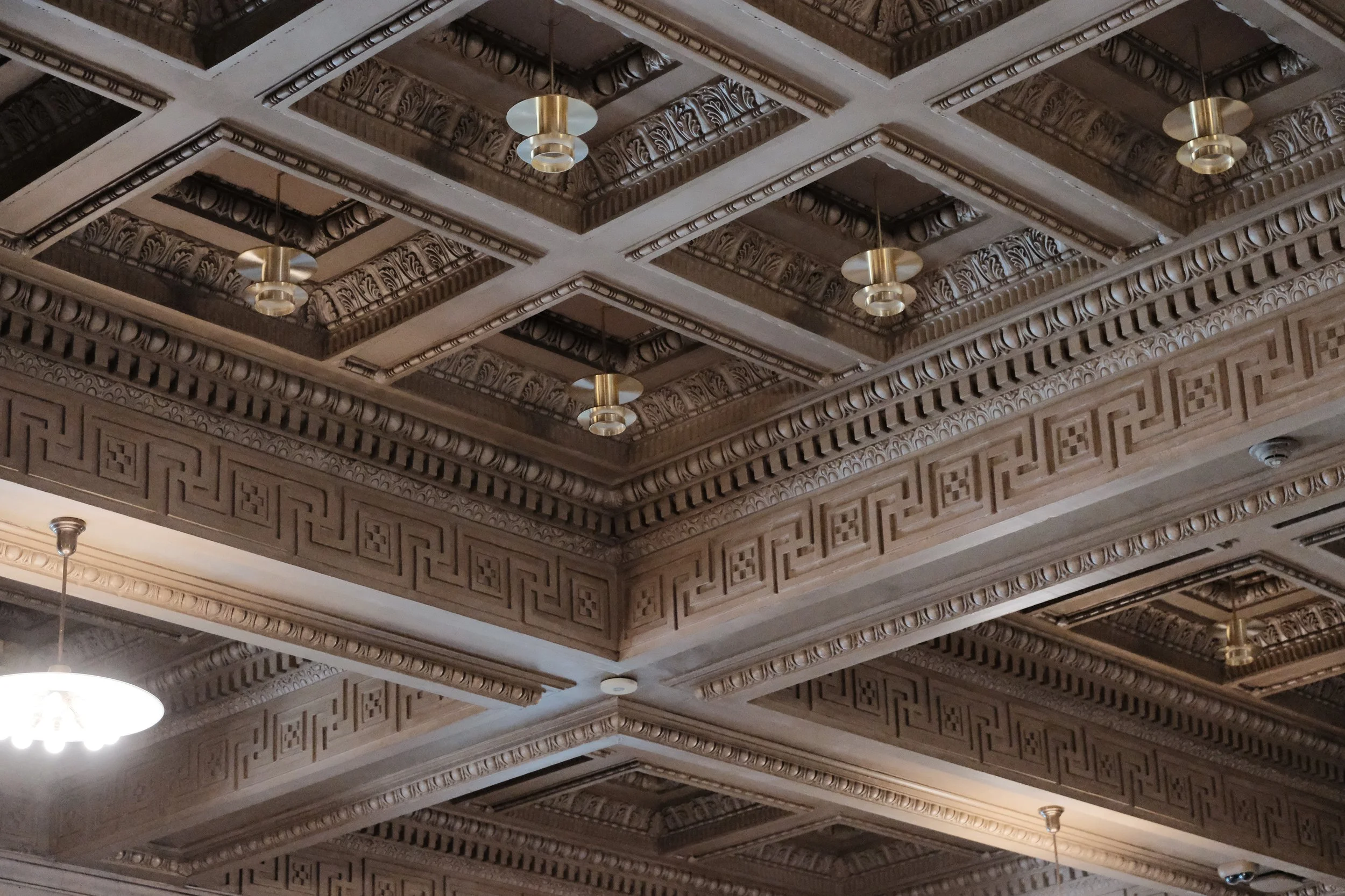 Ornate ceiling with patterned wood panels, decorative molding, and hanging light fixtures in a historic building.
