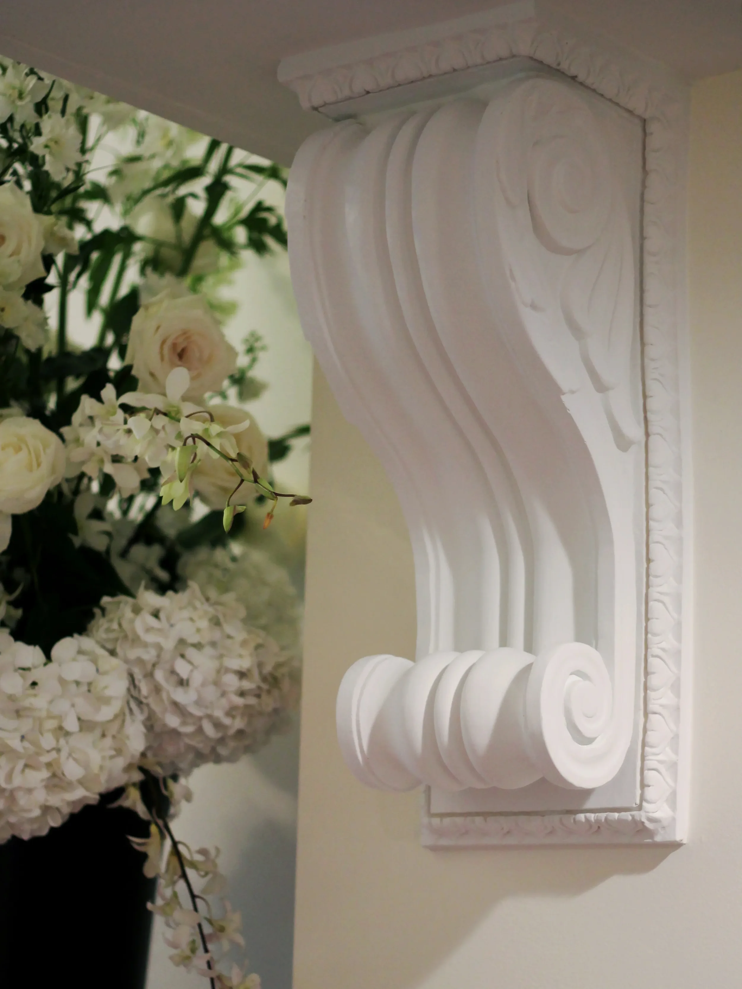 Close-up of a white decorative wall corbel with floral and scroll design details, mounted on a wall beside a bouquet of white flowers.