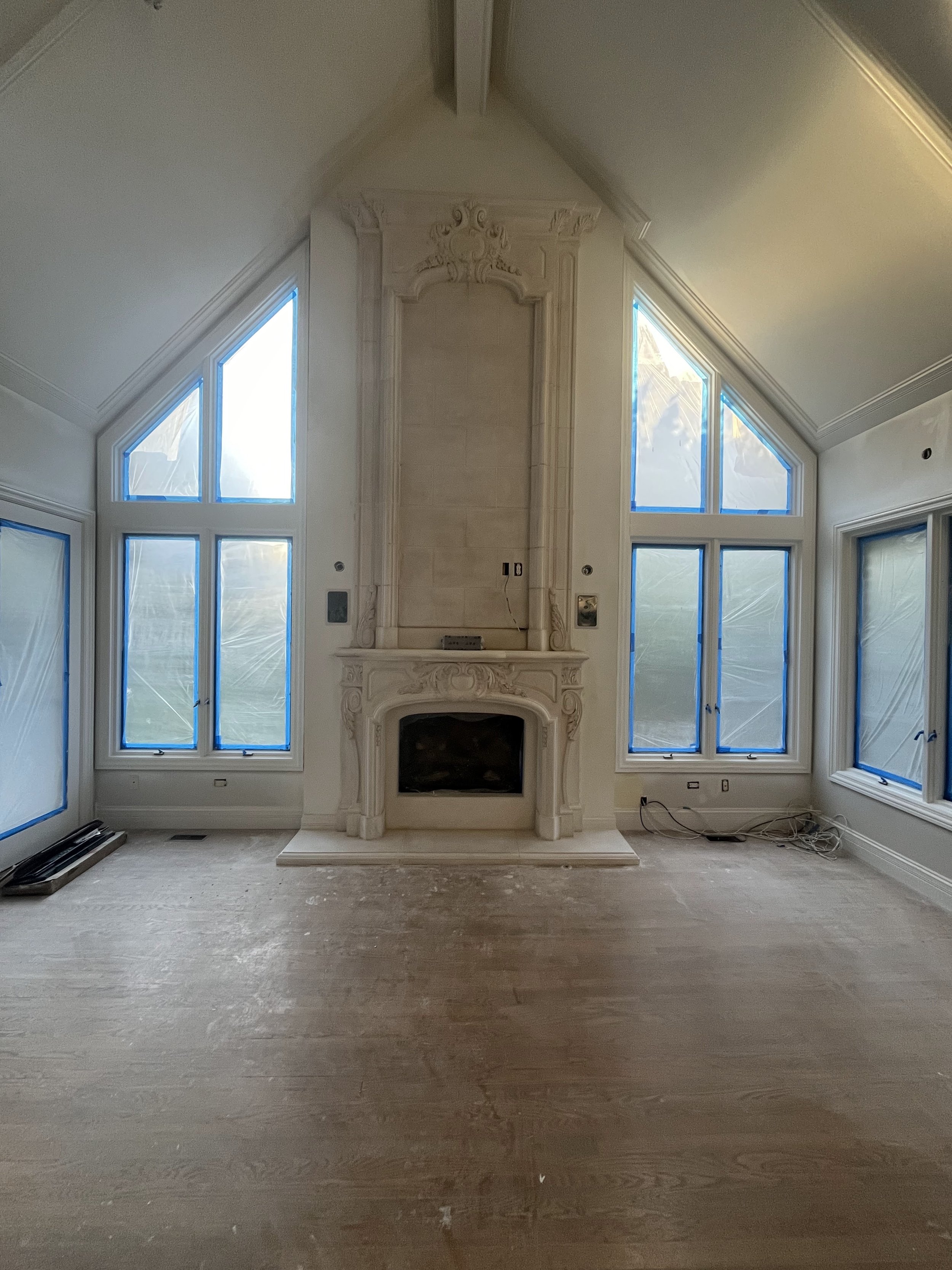 Living room under renovation with large windows covered in blue painter's tape and a decorative fireplace with a missing mantel, no furniture, and construction materials on the floor.