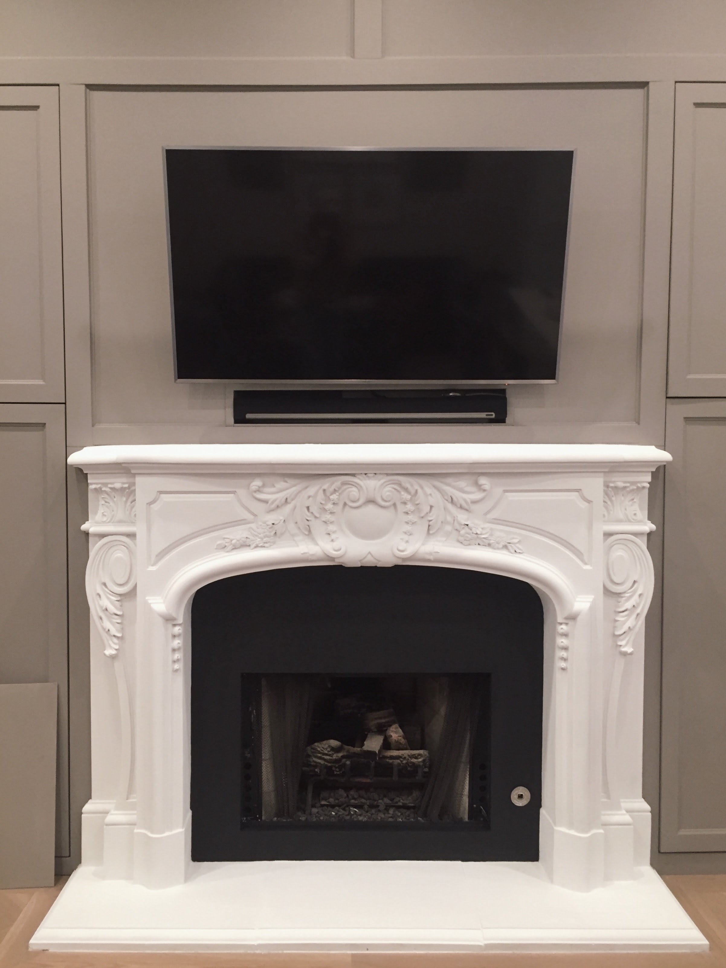 A flat-screen TV mounted above an ornate white fireplace with intricate carvings, set against a light gray wall with built-in cabinetry.