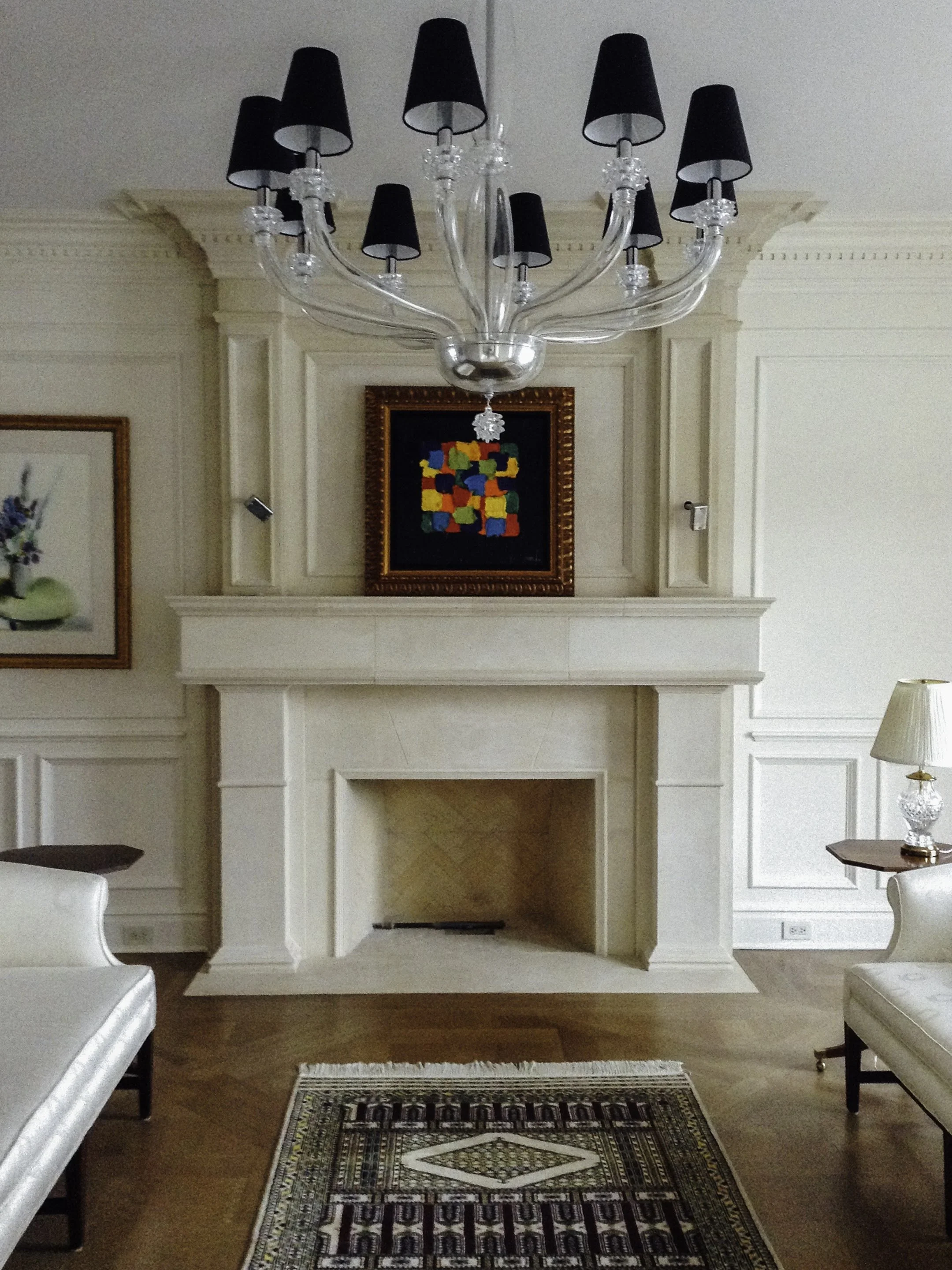 A living room with a white fireplace, hardwood flooring, and white paneled walls. There are two white armchairs, and a large glass chandelier with black lampshades hanging from the ceiling. A colorful abstract painting is above the fireplace.