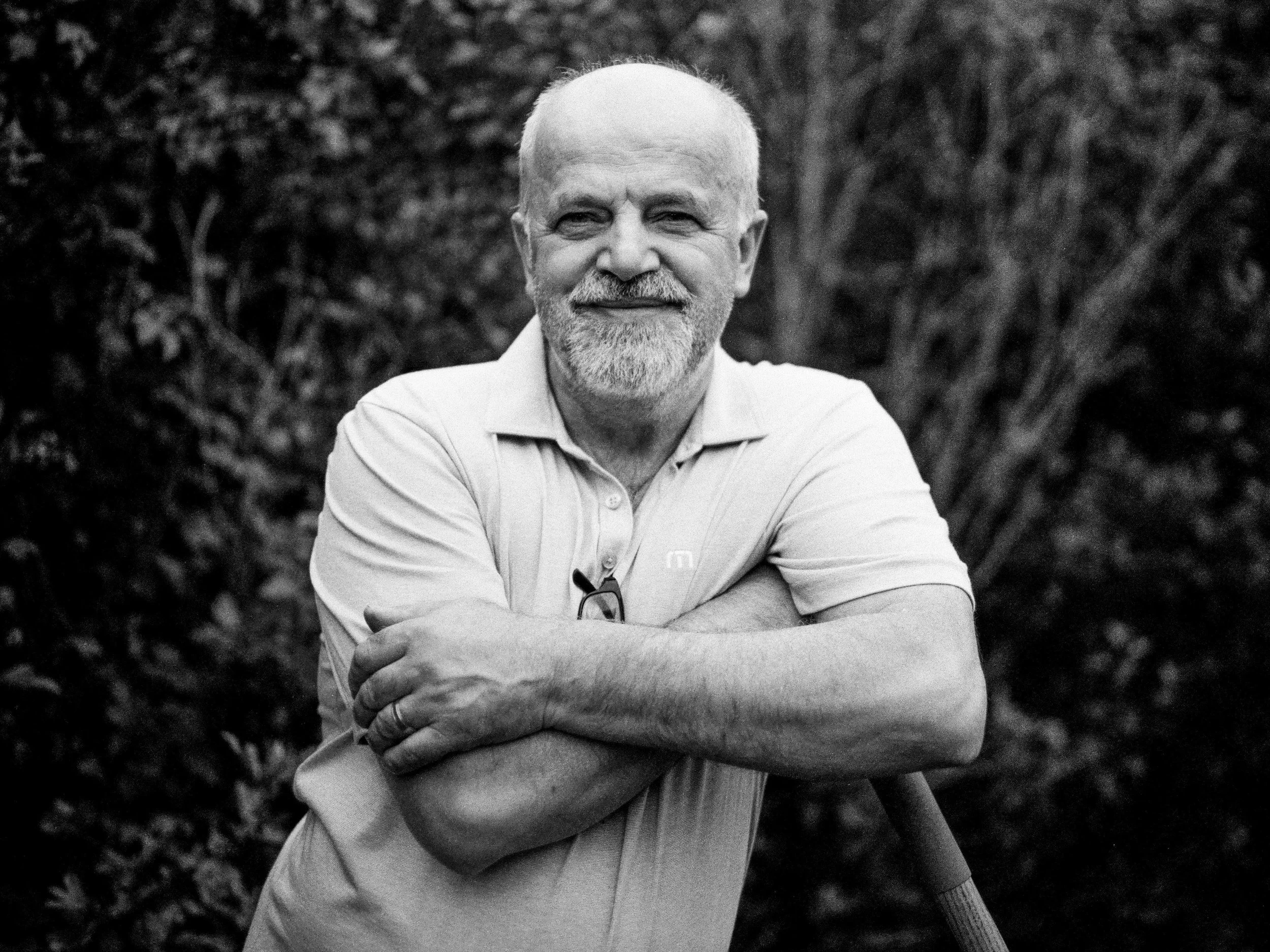A smiling elderly man with a beard and glasses resting on his shirt, standing outdoors with arms crossed, surrounded by bushes.