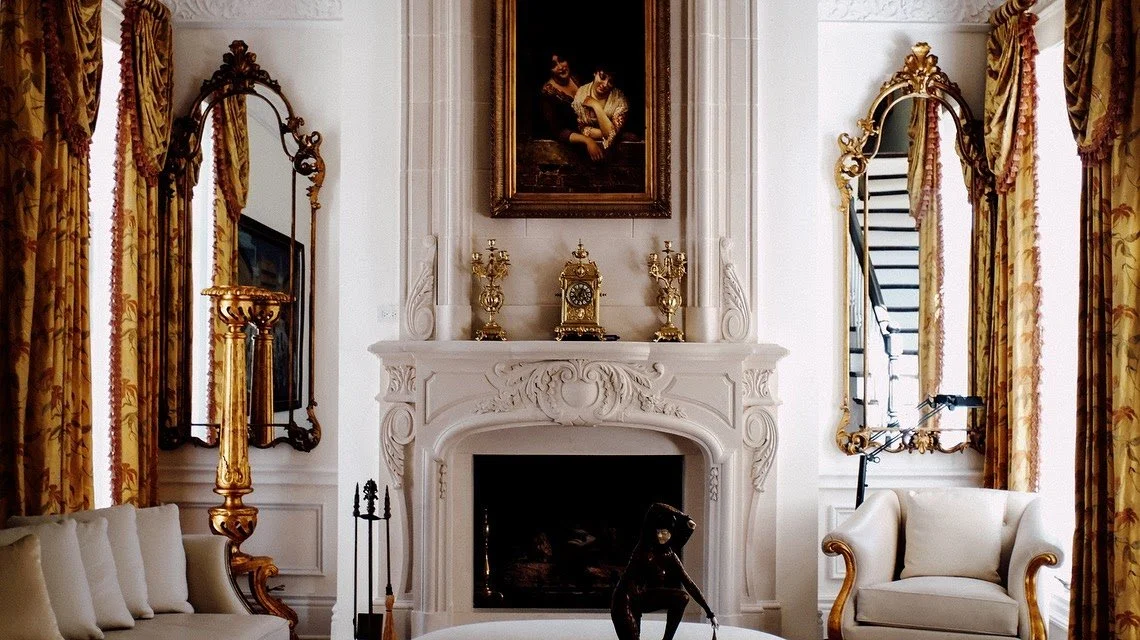 Elegant living room with ornate white fireplace, gold decorative items on mantel, gilded mirrors, and floral curtains.