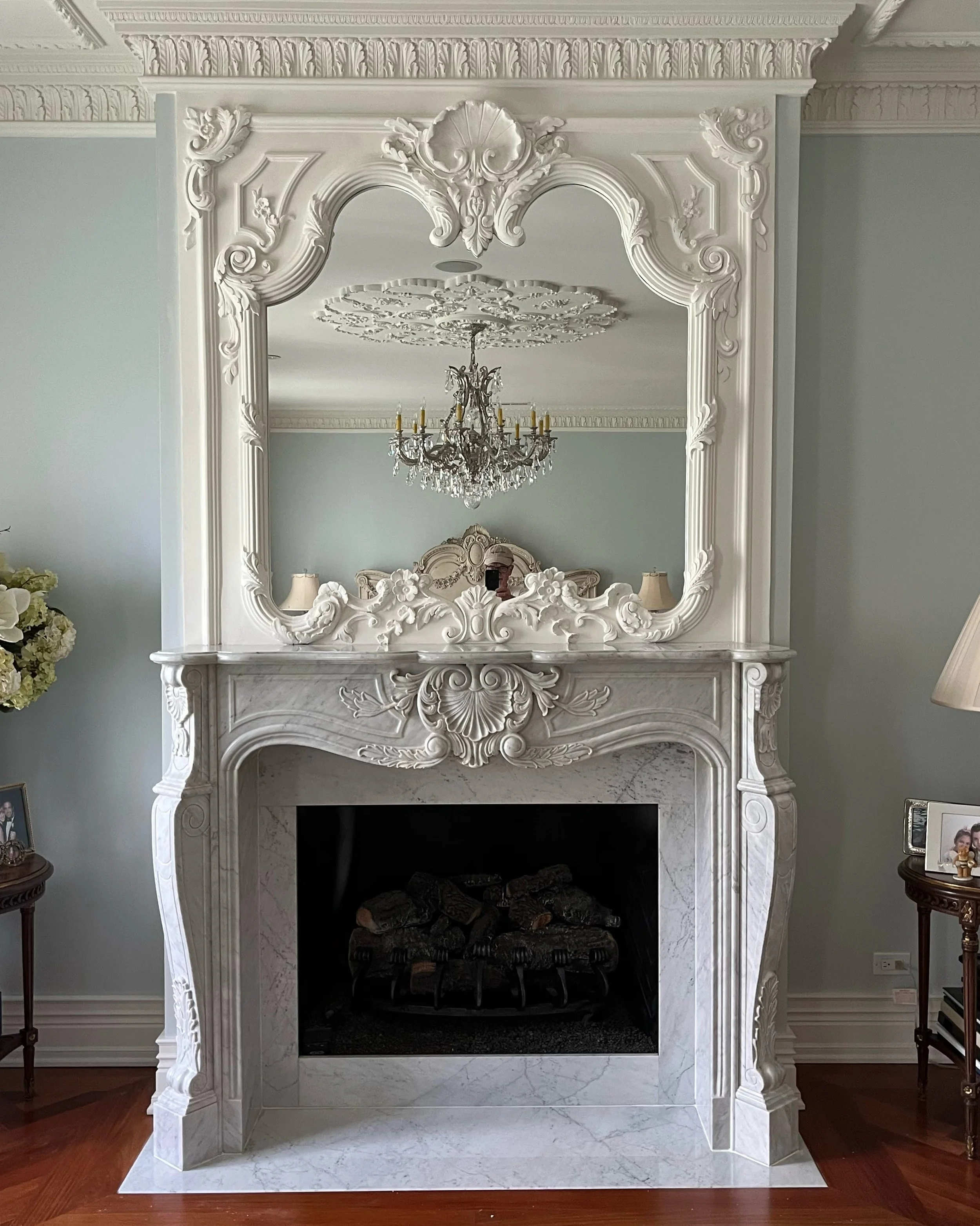Elegant white marble fireplace with ornate carvings and a large mirror above it, reflecting a chandelier and decorative ceiling details in a luxurious living room.