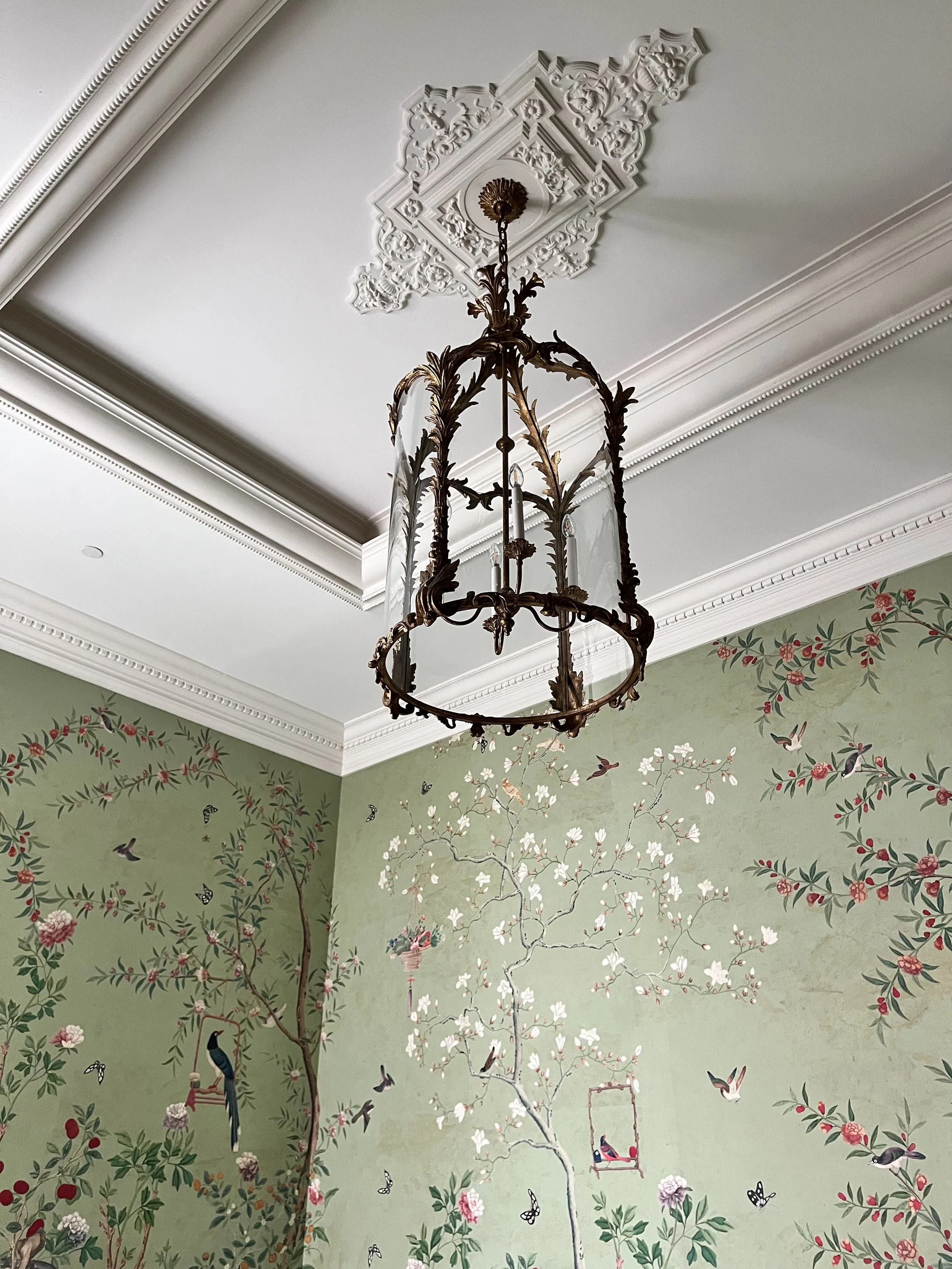 A decorative ceiling with a bronze chandelier hanging from an ornate ceiling medallion, and a painted wall featuring a nature scene with trees, flowers, birds, and butterflies.