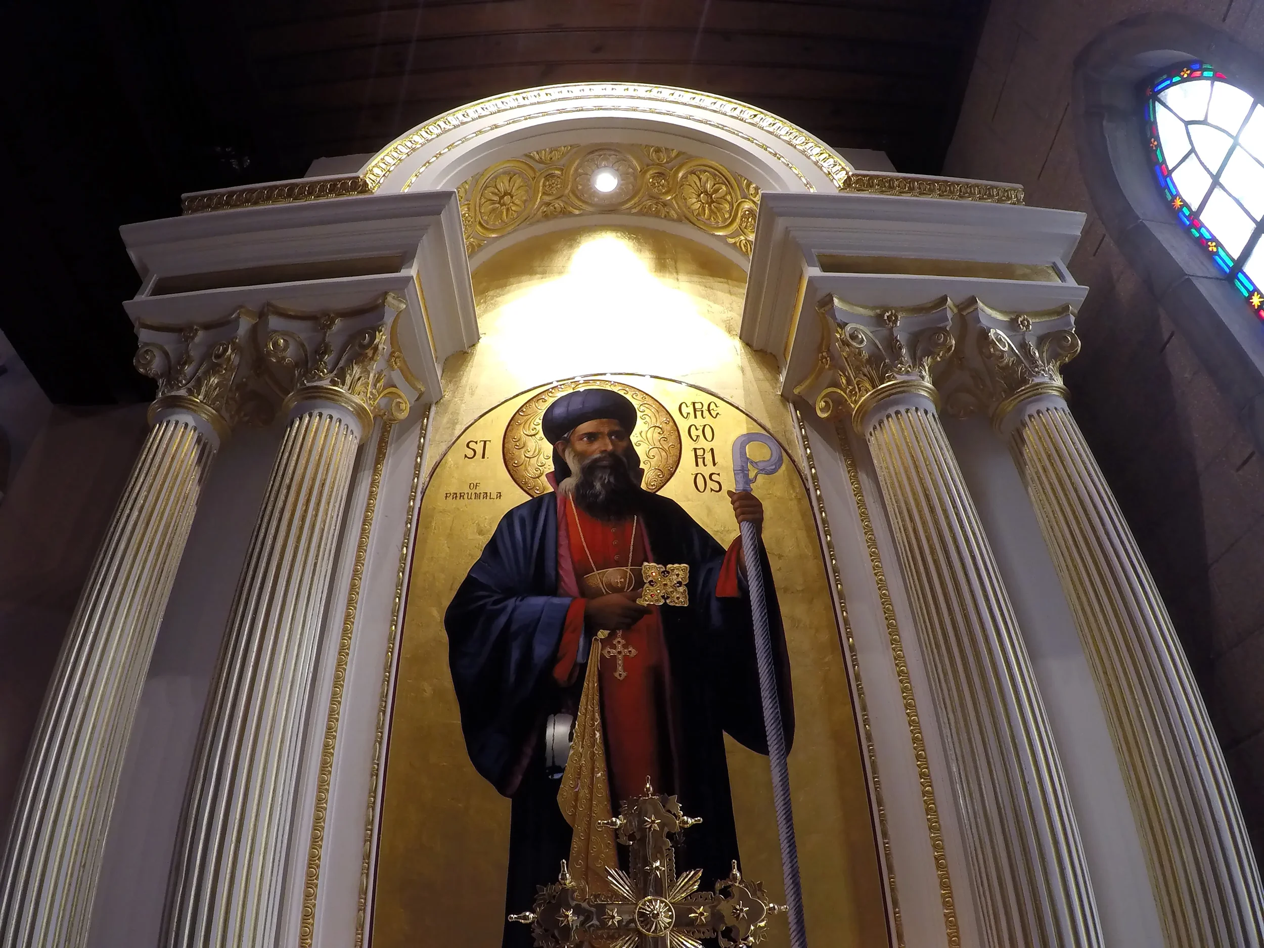 Religious statue of a saint in a church, surrounded by ornate gold and white columns with a stained glass window nearby.