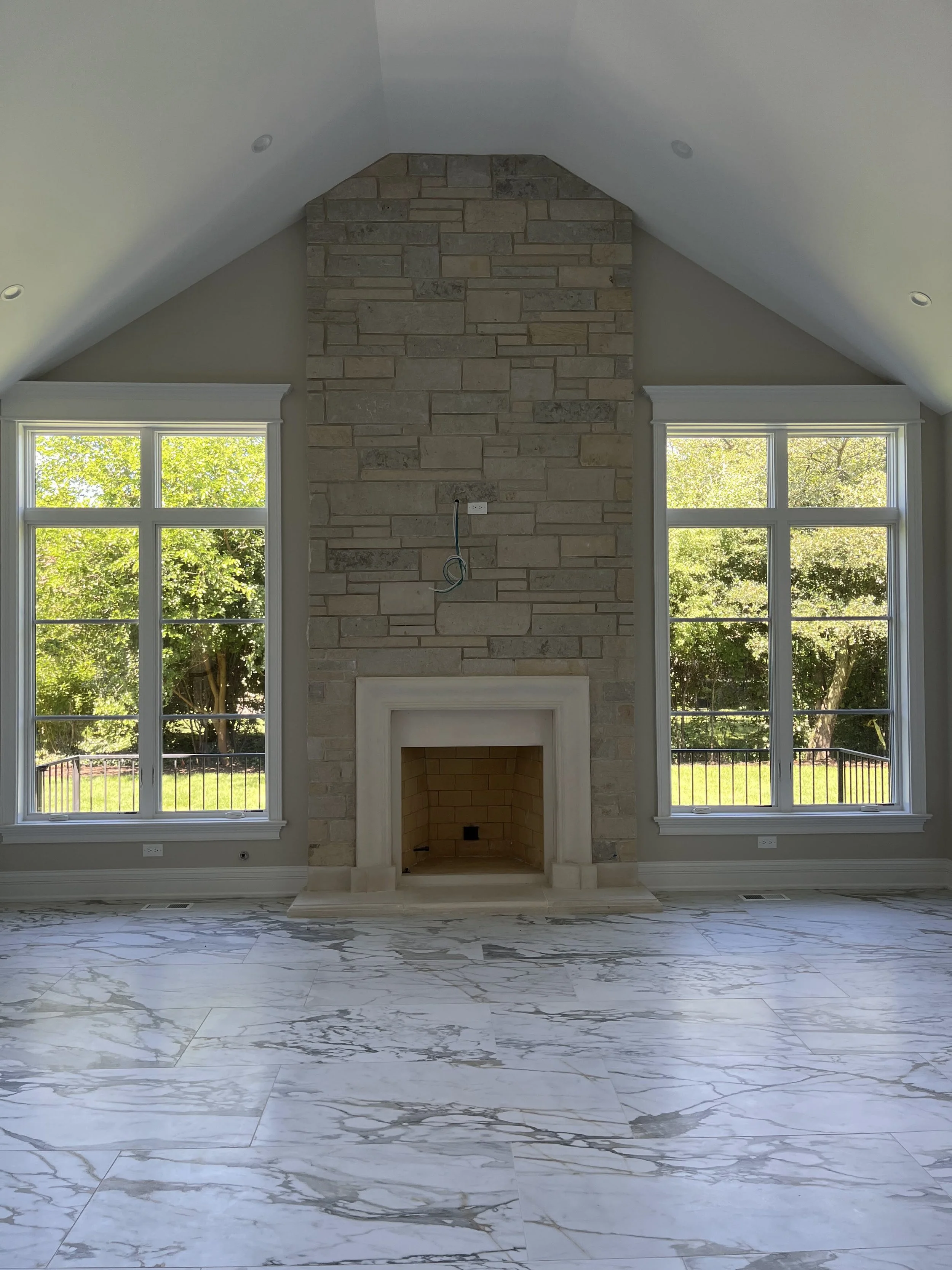 Empty living room with a stone fireplace and two large windows overlooking a backyard with green trees.
