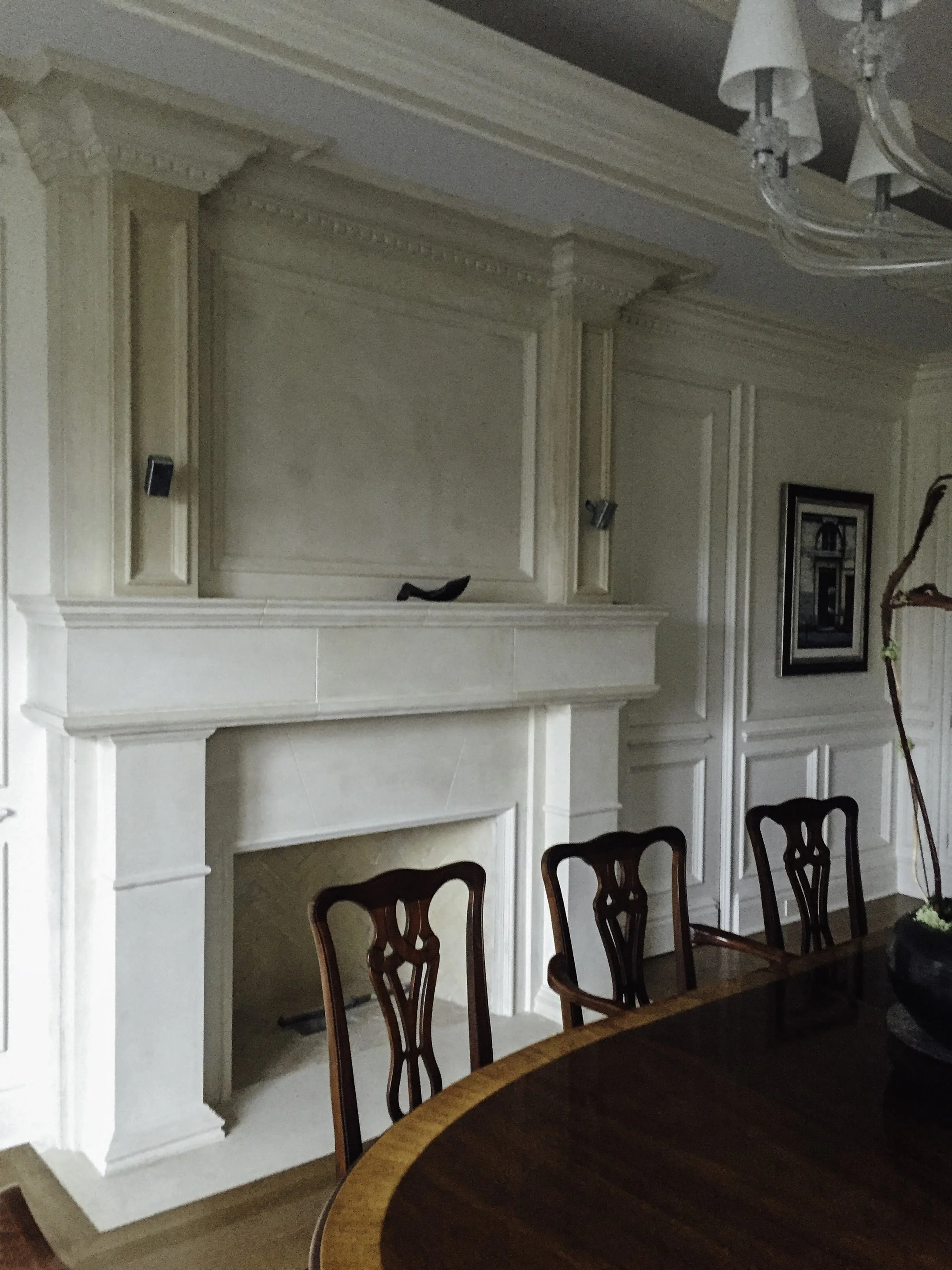 Interior of a dining room with a white ornate fireplace and paneling, dark wooden chairs around a dark wooden table, and a chandelier hanging from the ceiling.
