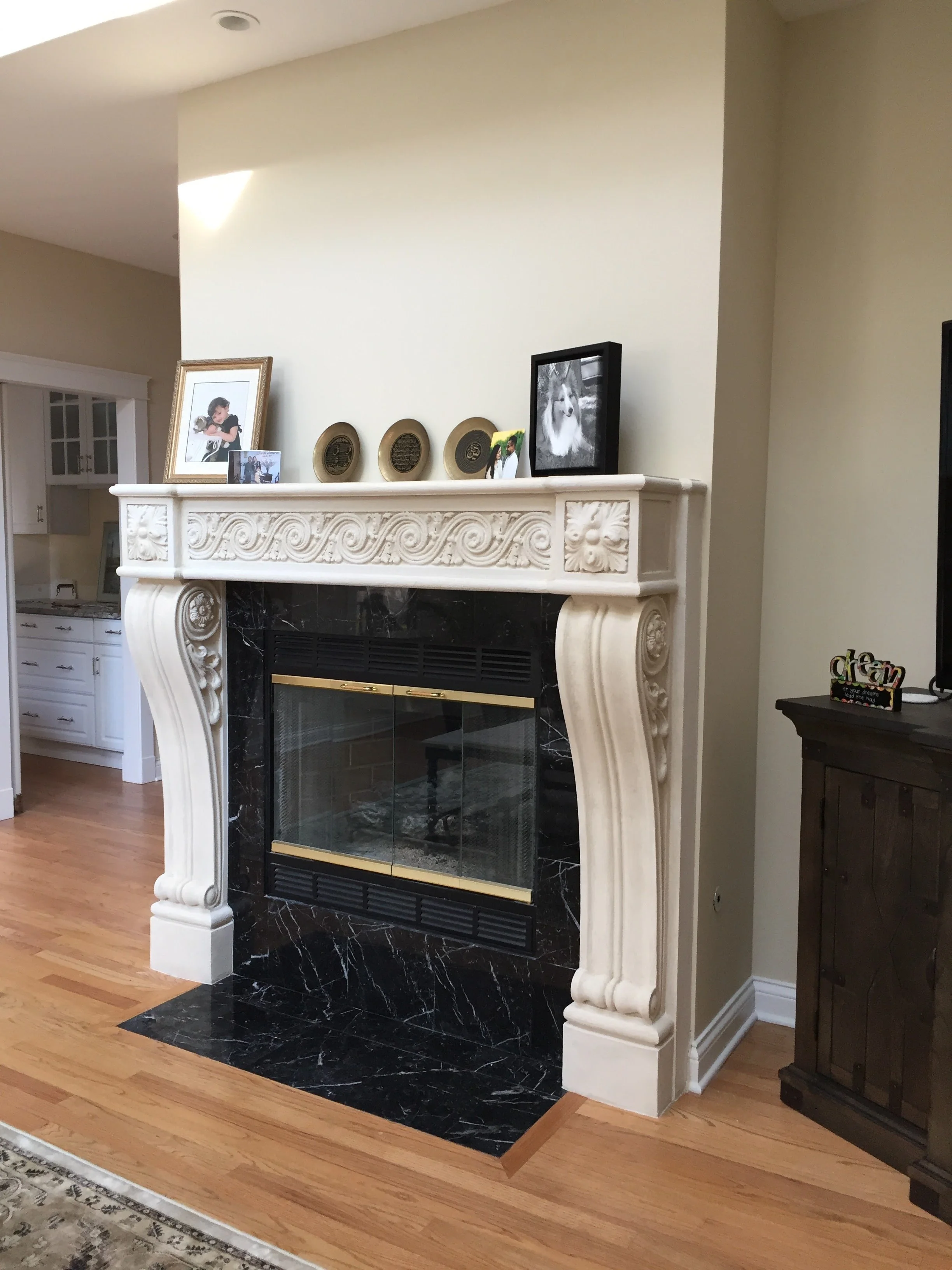 A living room fireplace with ornate white carved mantel and black marble surround, decorated with framed photographs and decorative plates.