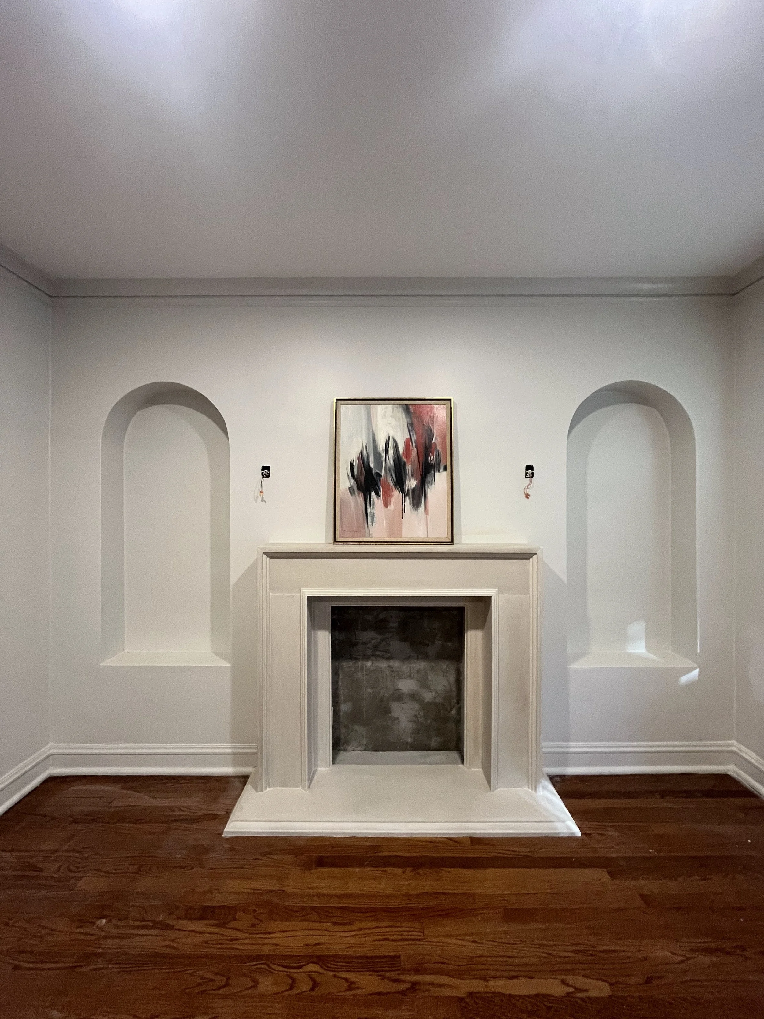 Empty living room with white walls, a white fireplace, a framed abstract painting above the fireplace, and recessed wall niches, hardwood floor.
