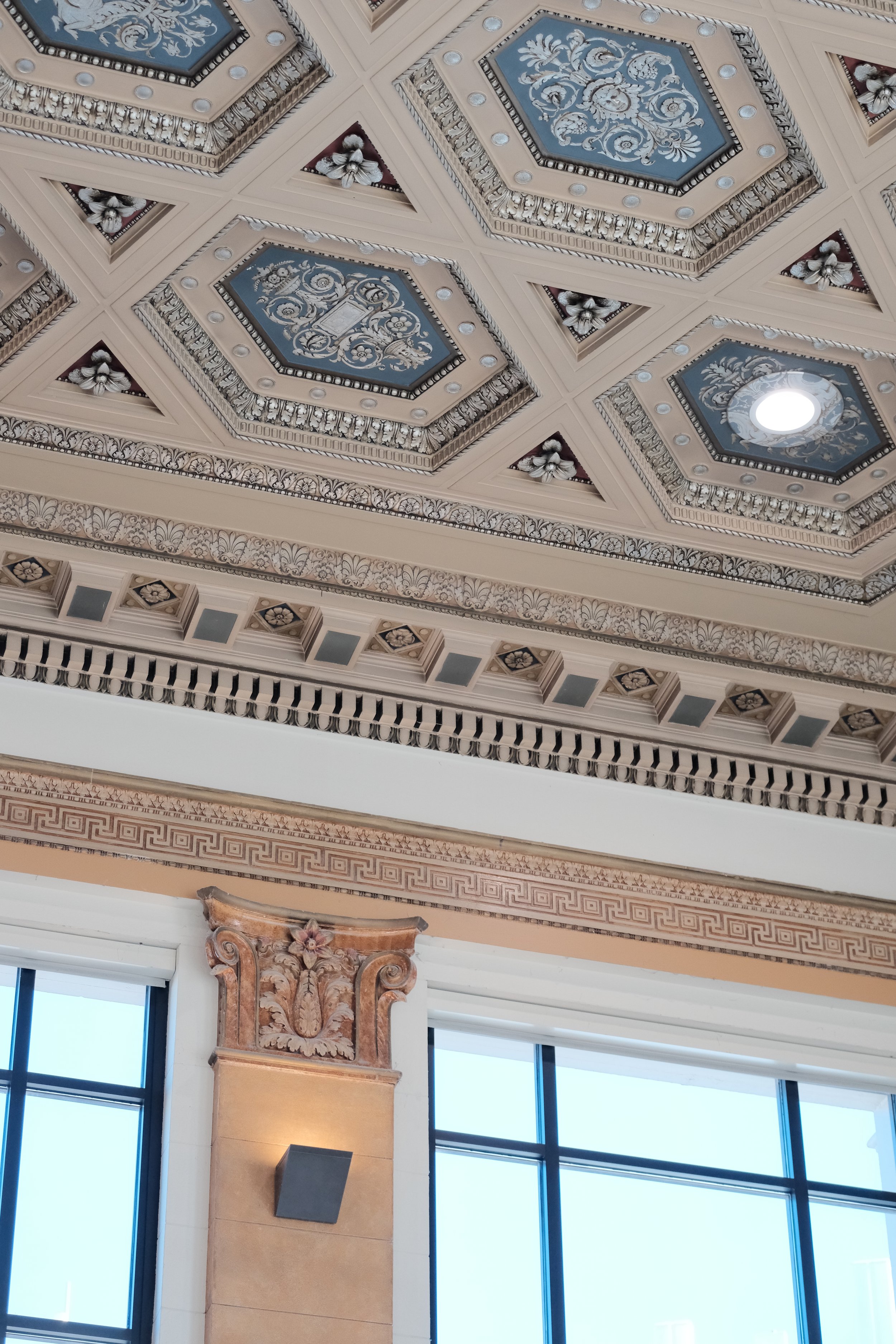Decorative ceiling with ornate moldings, painted panels, and a skylight, along with large windows and classical architectural elements.