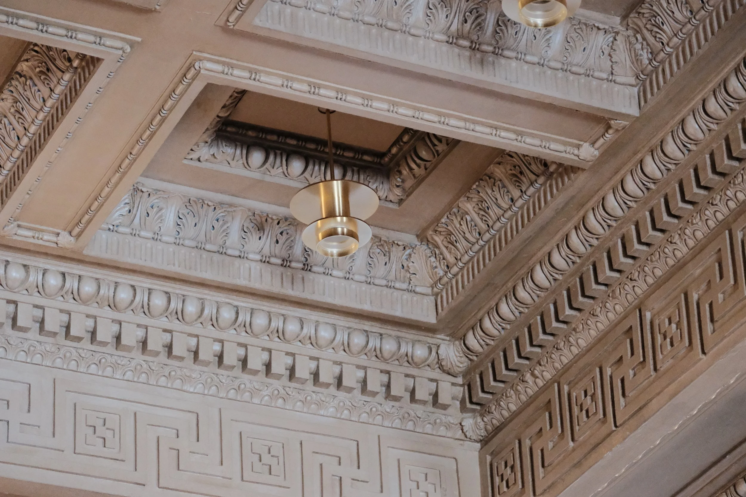 Ceiling with ornate decorative molding, gold-colored hanging light fixture, and intricate patterns.