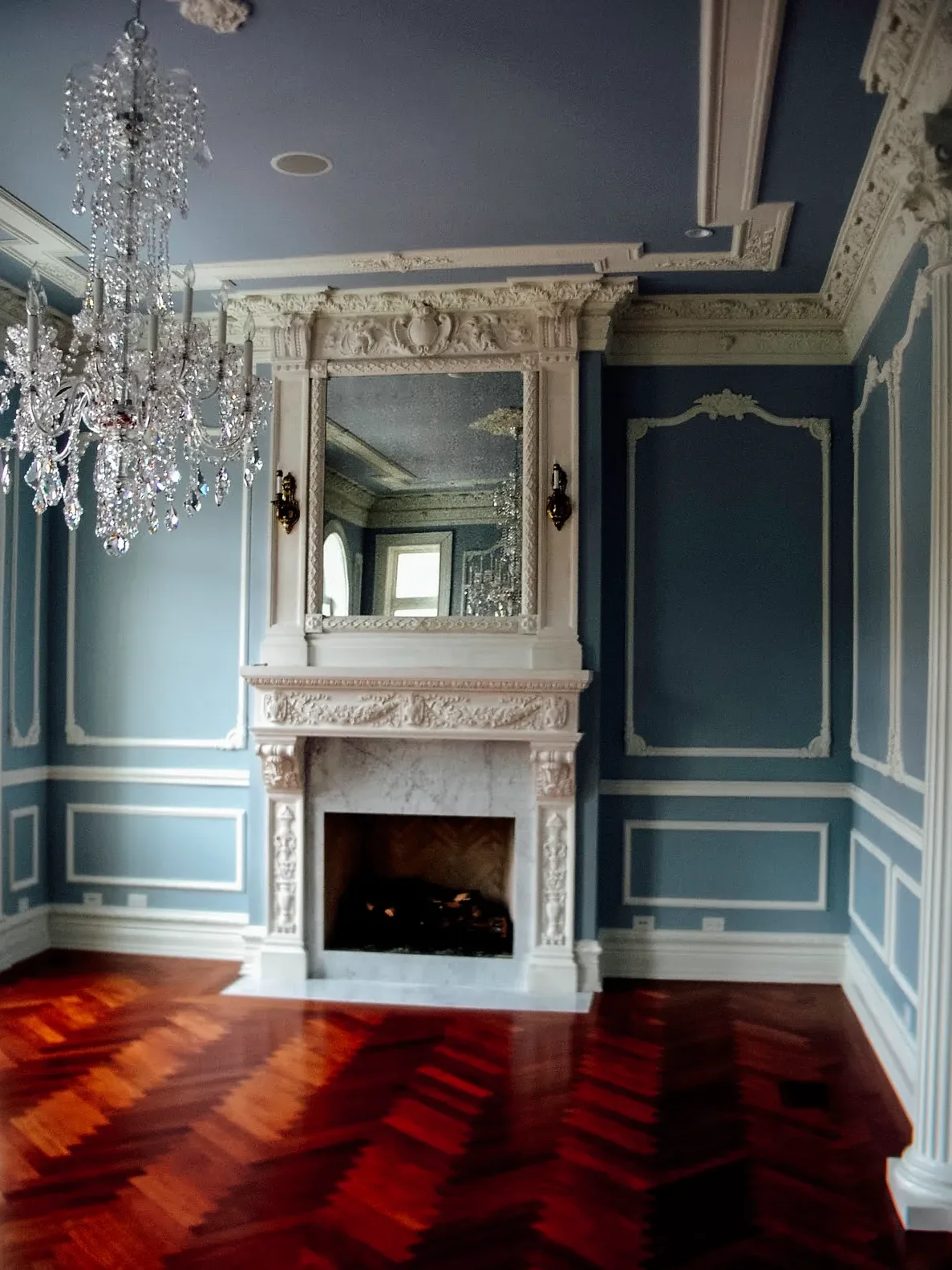 Elegant room with blue walls, detailed white crown molding, a marble fireplace, a large ornate mirror above the fireplace, a chandelier hanging from the ceiling, and hardwood floors.