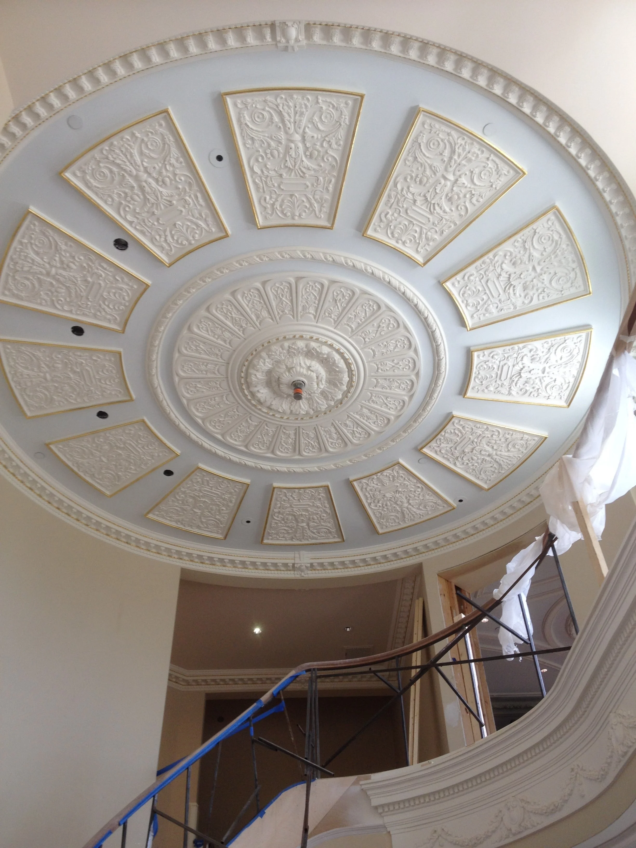 Ceiling with elaborate decorative plasterwork featuring a circular pattern with ornate panels and a central medallion, under renovation with scaffolding and construction materials.