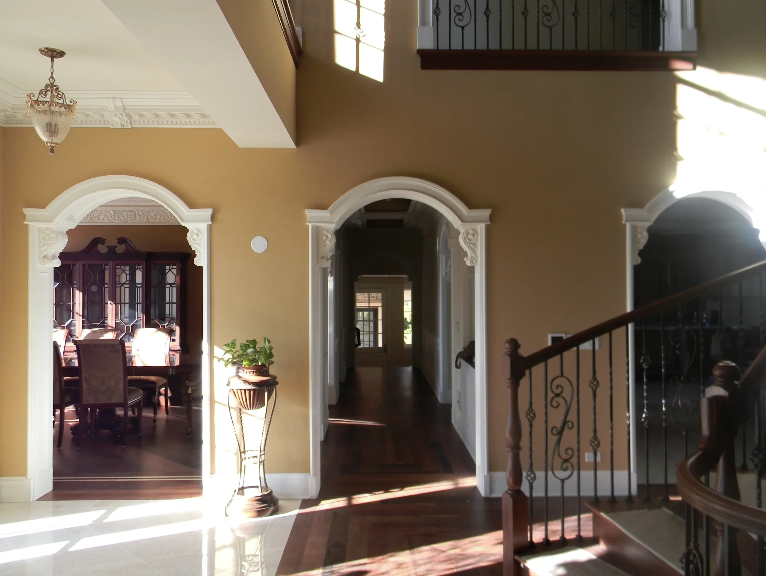 Interior view of a house showing a hallway with archways leading to a dining room with a wooden china cabinet. The hallway has hardwood floors, and there is a staircase with a wooden handrail on the right. Sunlight is streaming into the house, illumi