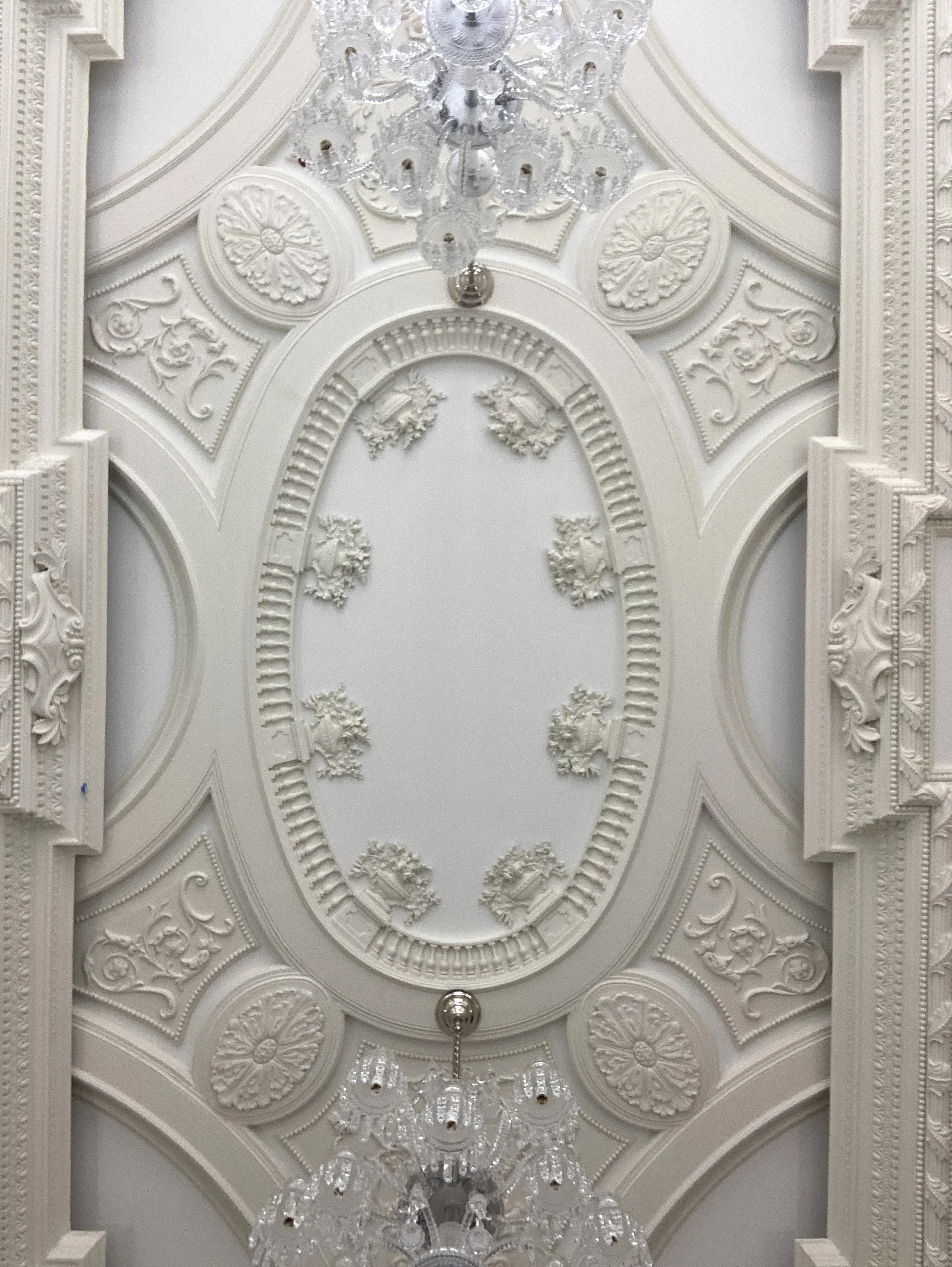 Ornate ceiling with intricate white moldings, floral decorations, and two elegant chandeliers.