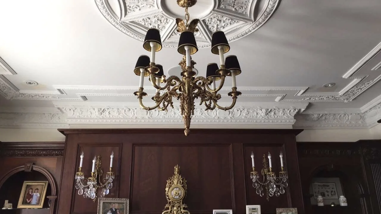Elegant living room with ornate white ceiling, large gold chandelier with black lampshades, dark wood paneling, and gold and crystal wall sconces.