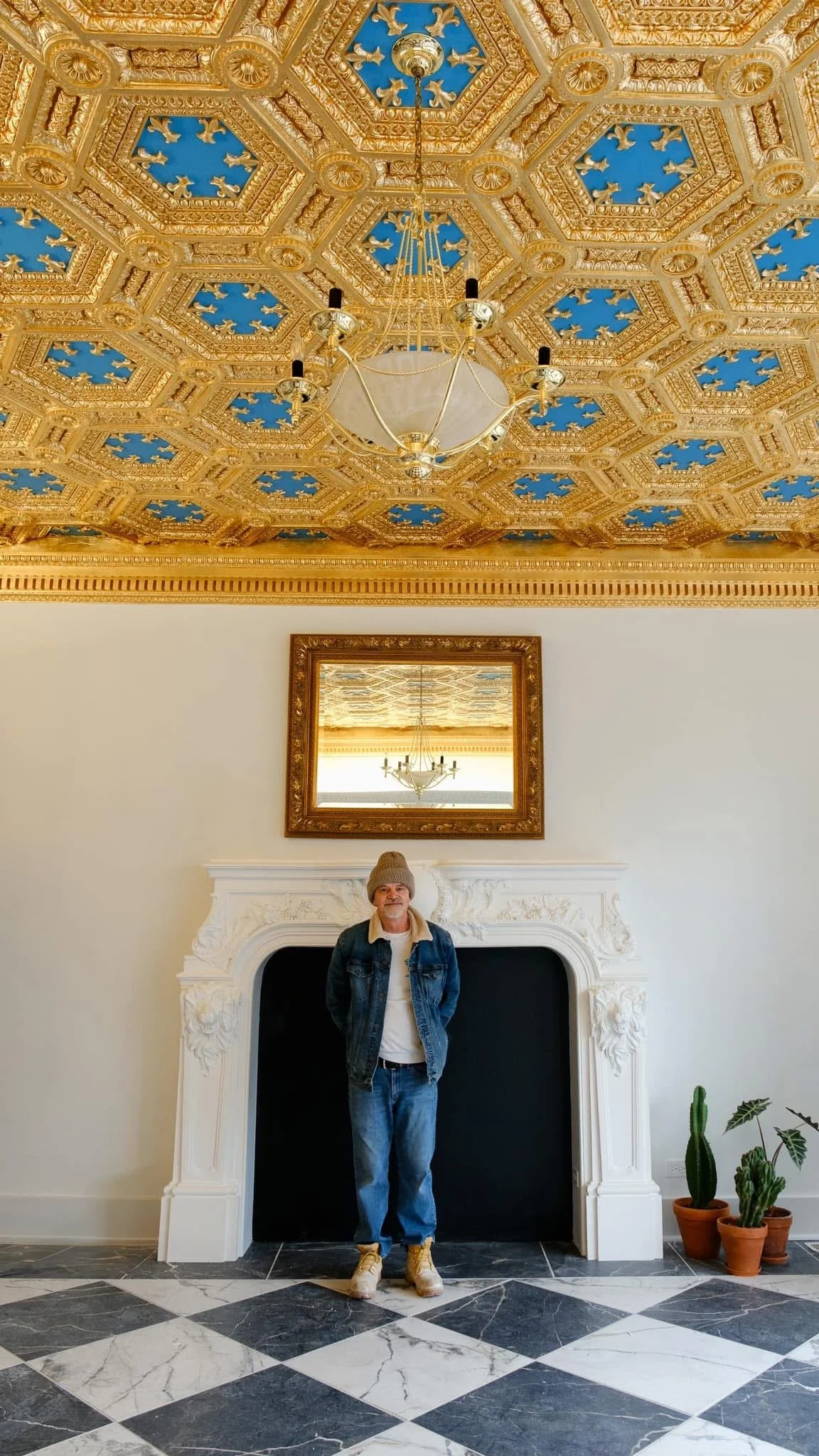 A man wearing a gray beanie, denim jacket, white shirt, and jeans stands in front of a ornate white fireplace with black interior, in a room with a gold decorated ceiling and a chandelier reflected in a mirror above the fireplace. Potted plants are o