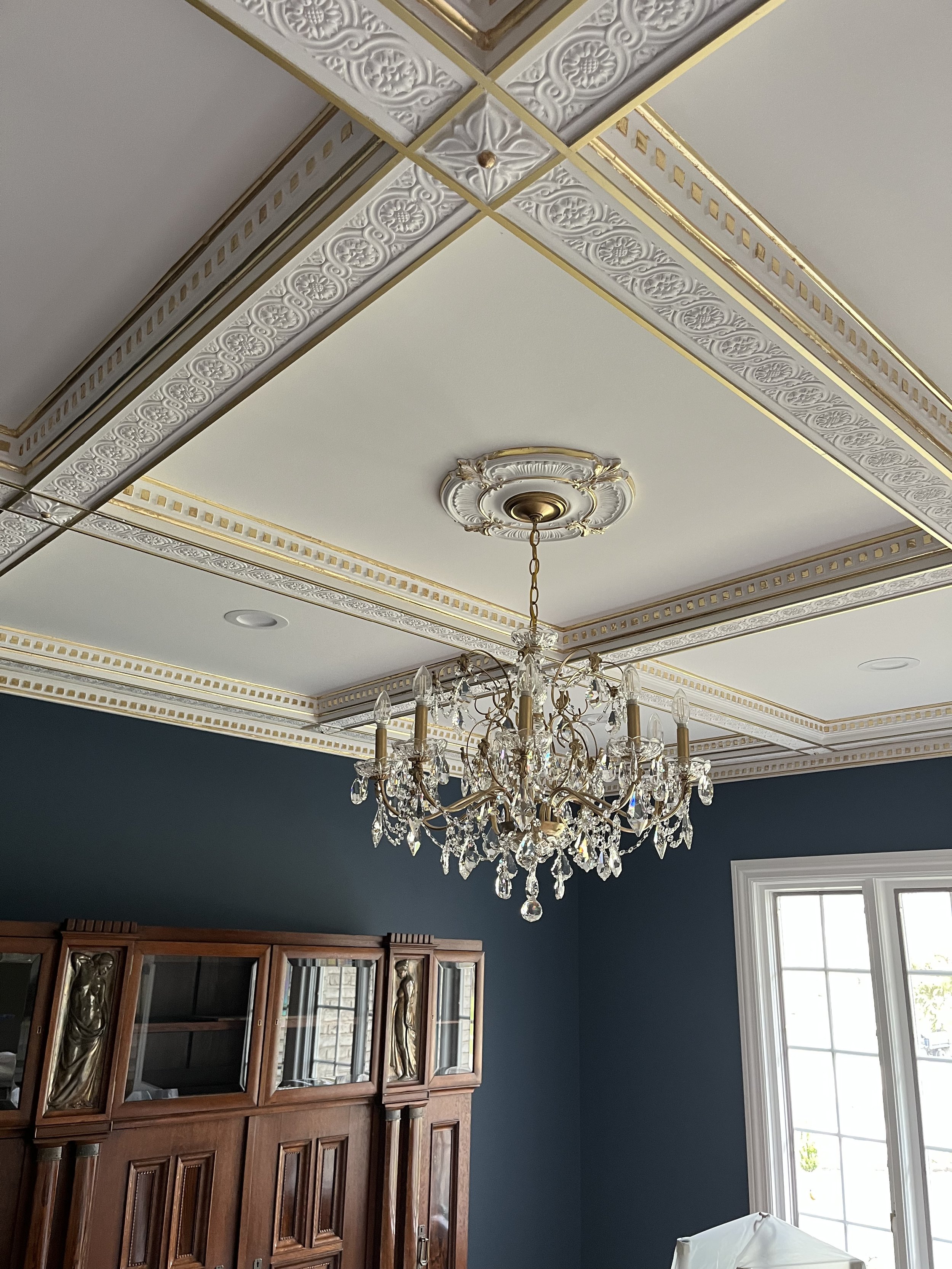Interior room featuring an ornate white ceiling with gold accents, a crystal chandelier hanging from the center, a dark green wall, a large window, and a wooden display cabinet with glass doors.