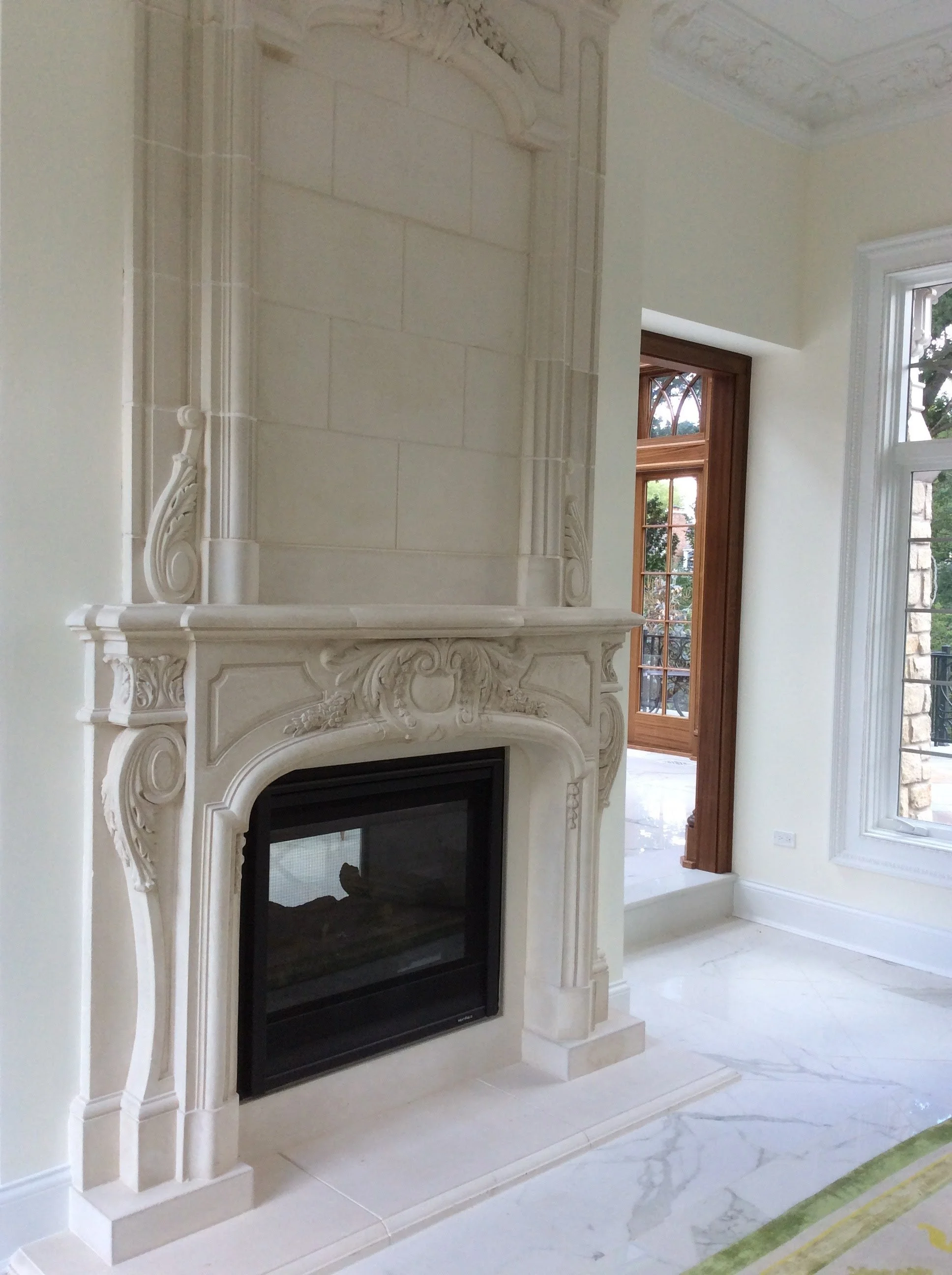 Beautiful living room with ornate white stone fireplace, large windows, and hardwood door leading outside.