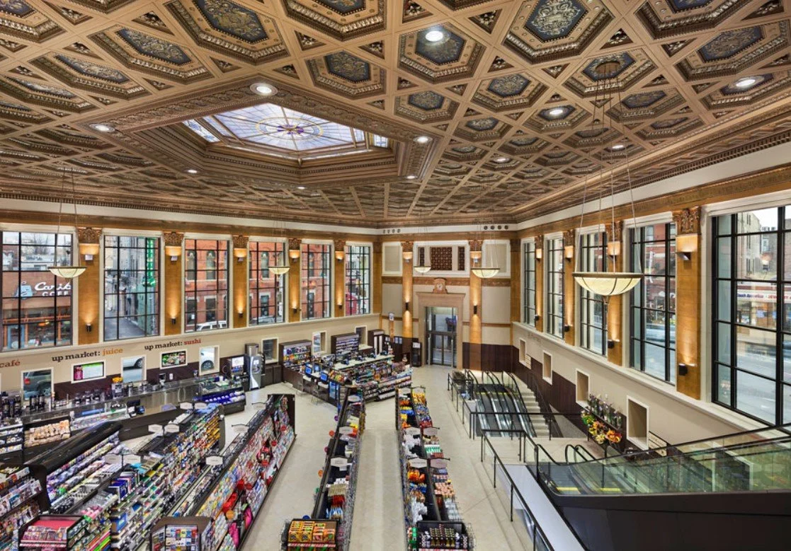 Interior of a grocery store with large windows, decorative ceiling, and aisles of shelves filled with various products.