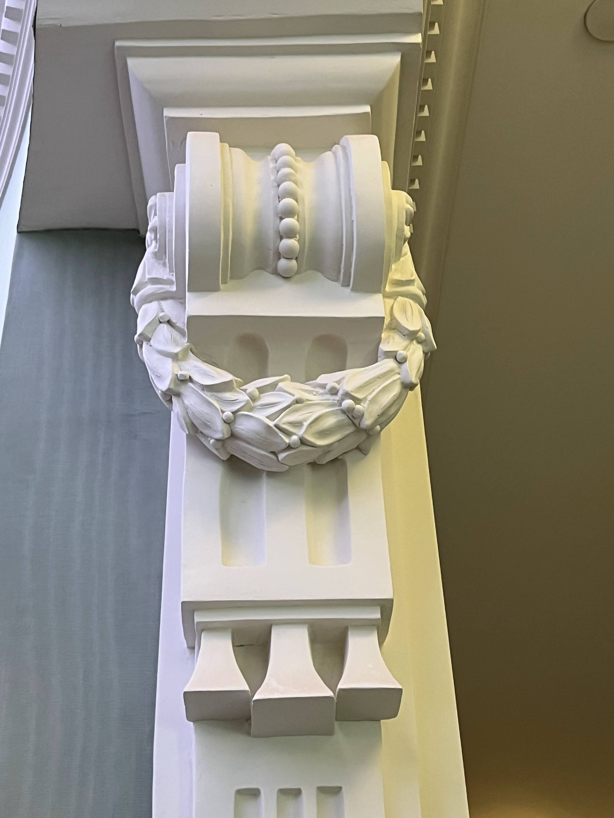 A white decorative architectural element with a semicircular design, adorned with bead-like ornaments, situated on a grey surface against a beige wall.