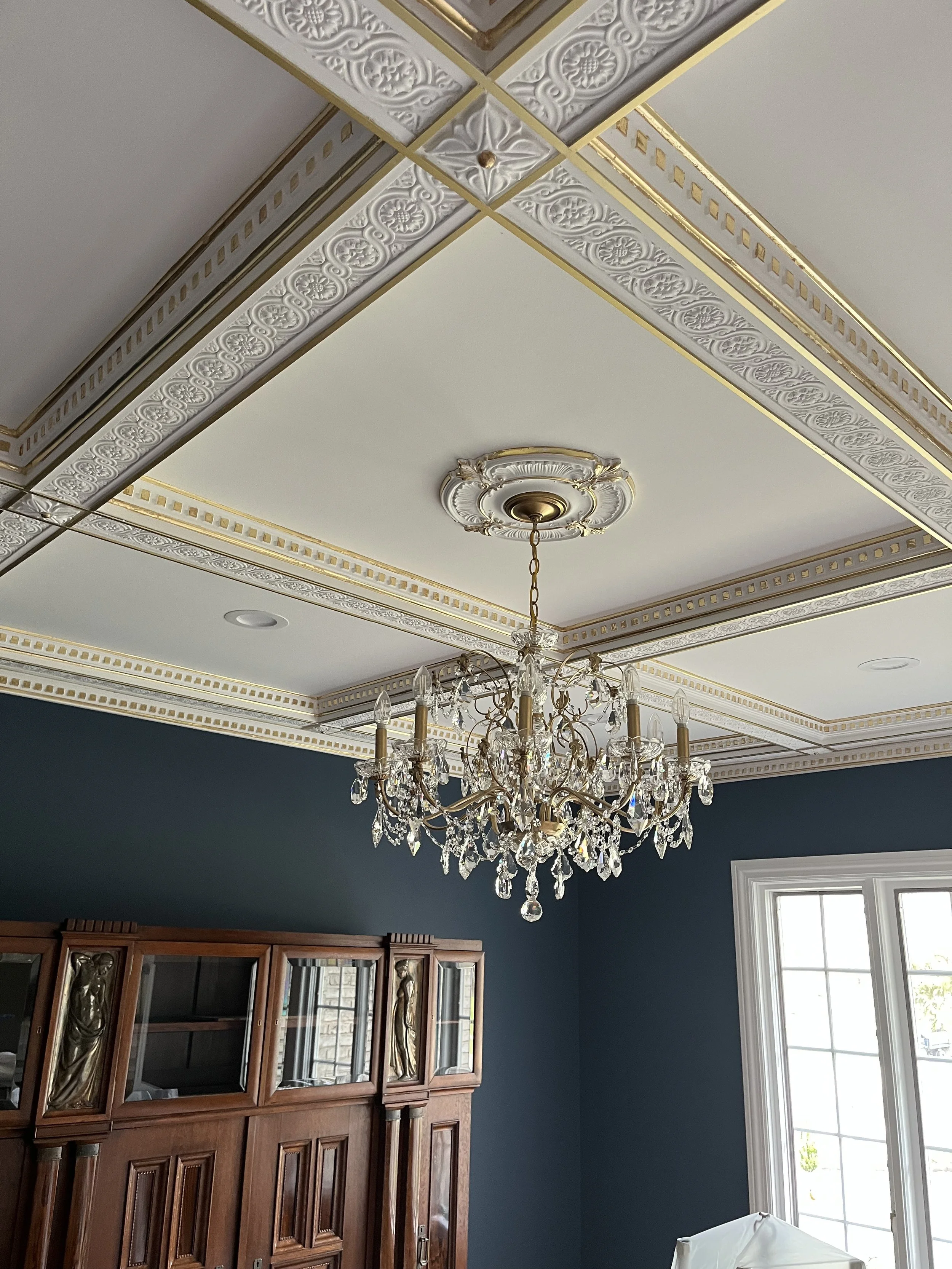 Interior room with a decorative ceiling, ornate chandelier, dark blue wall, wooden cabinet, and a window with white trim.