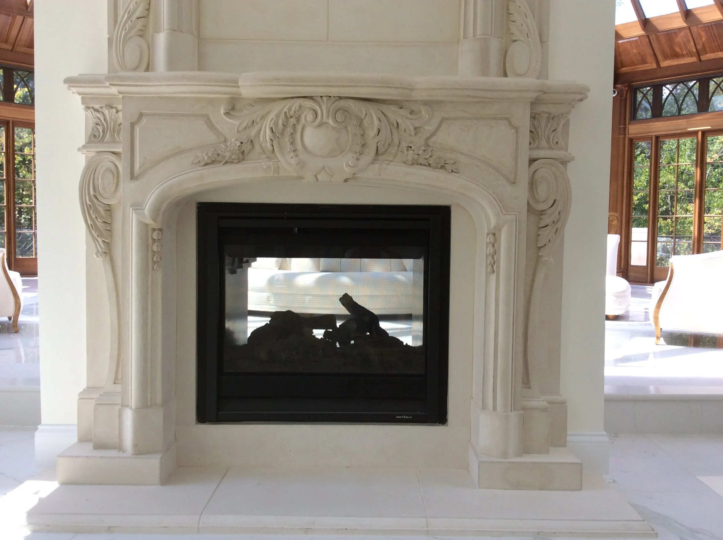 Ornate white marble fireplace with decorative carvings in a living room with large windows and sunlight.