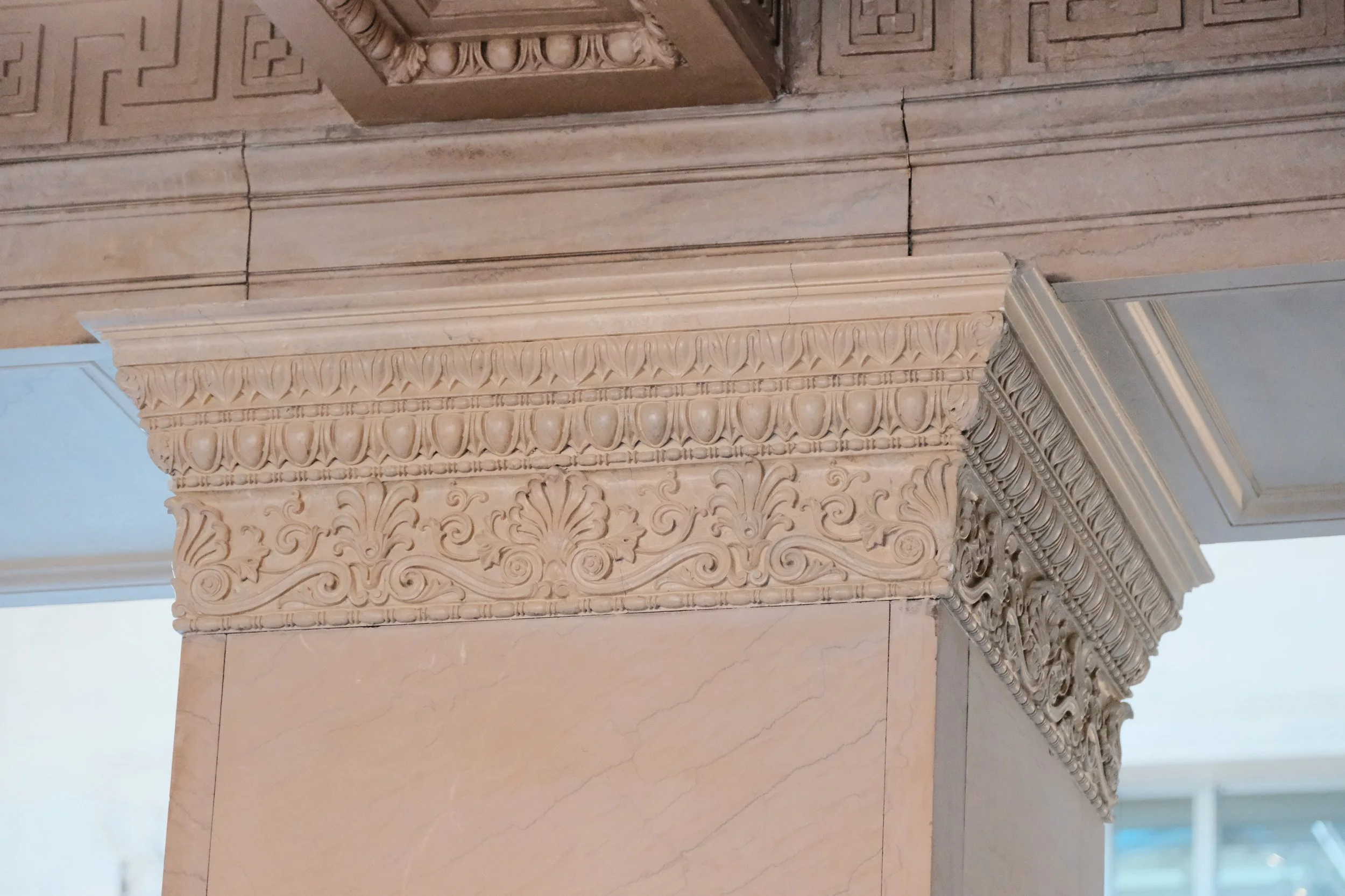 Close-up of ornate architectural detailing on a beige marble column and wall, featuring carved floral and scroll motifs.