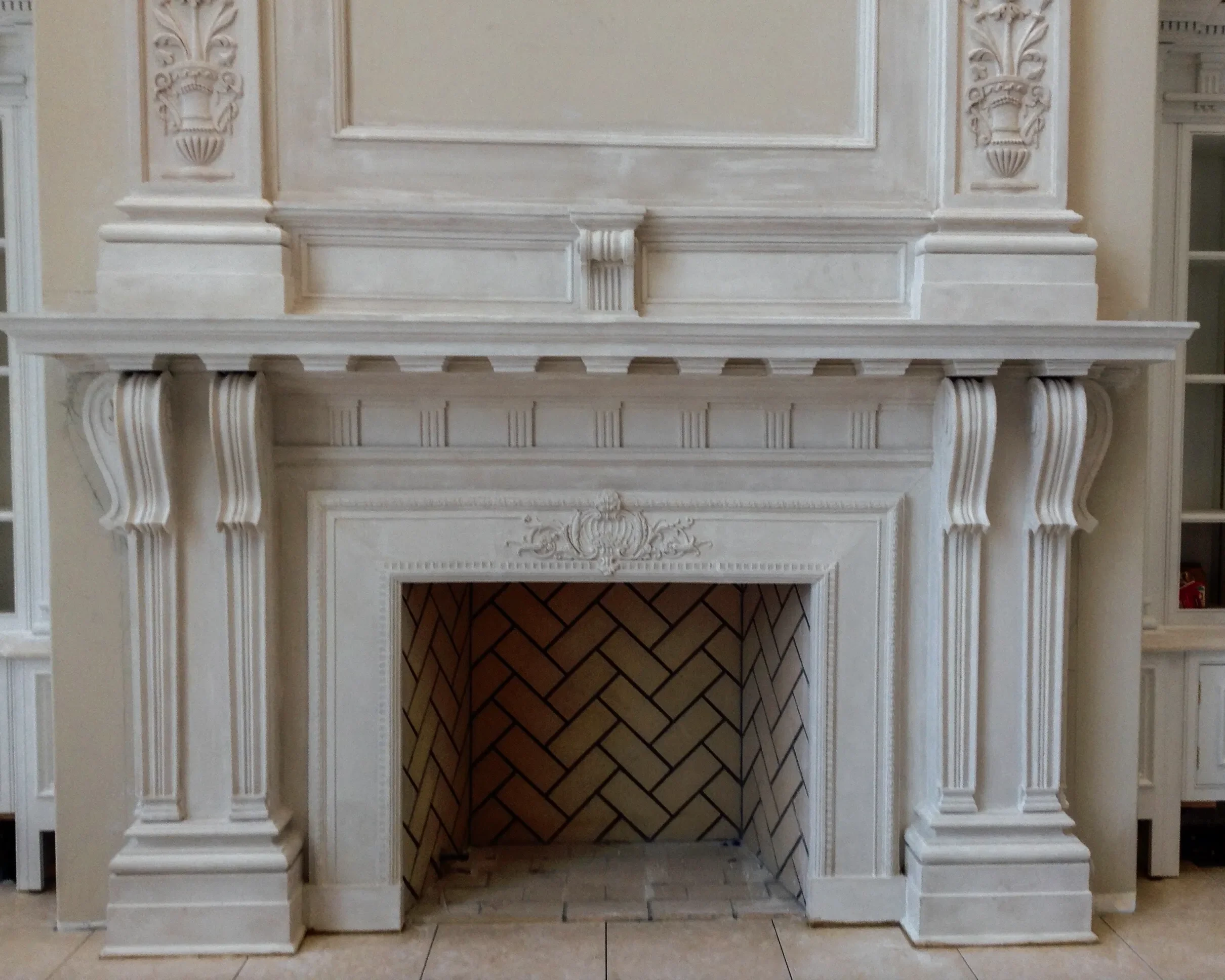 Elegant white stone fireplace with intricate carvings, columns, and a decorative mantel, with a tiled interior in a well-lit room.