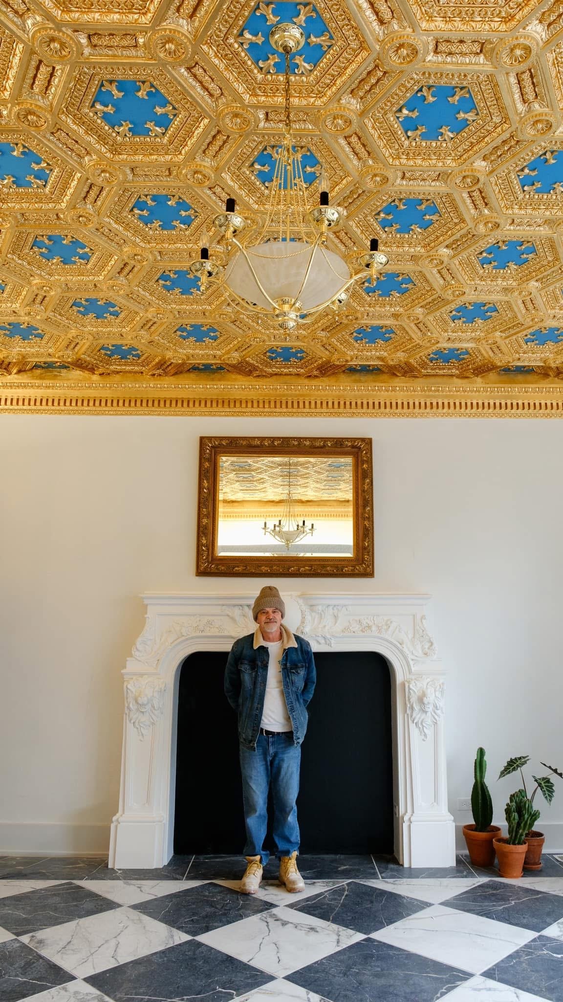 A man standing in front of a decorative fireplace with an ornate white mantel and a mirror above, inside a room with a highly decorative gold ceiling featuring intricate patterns and a large chandelier. There are potted cacti on the right side of the fireplace, and the floor is a black and white checkered tile.