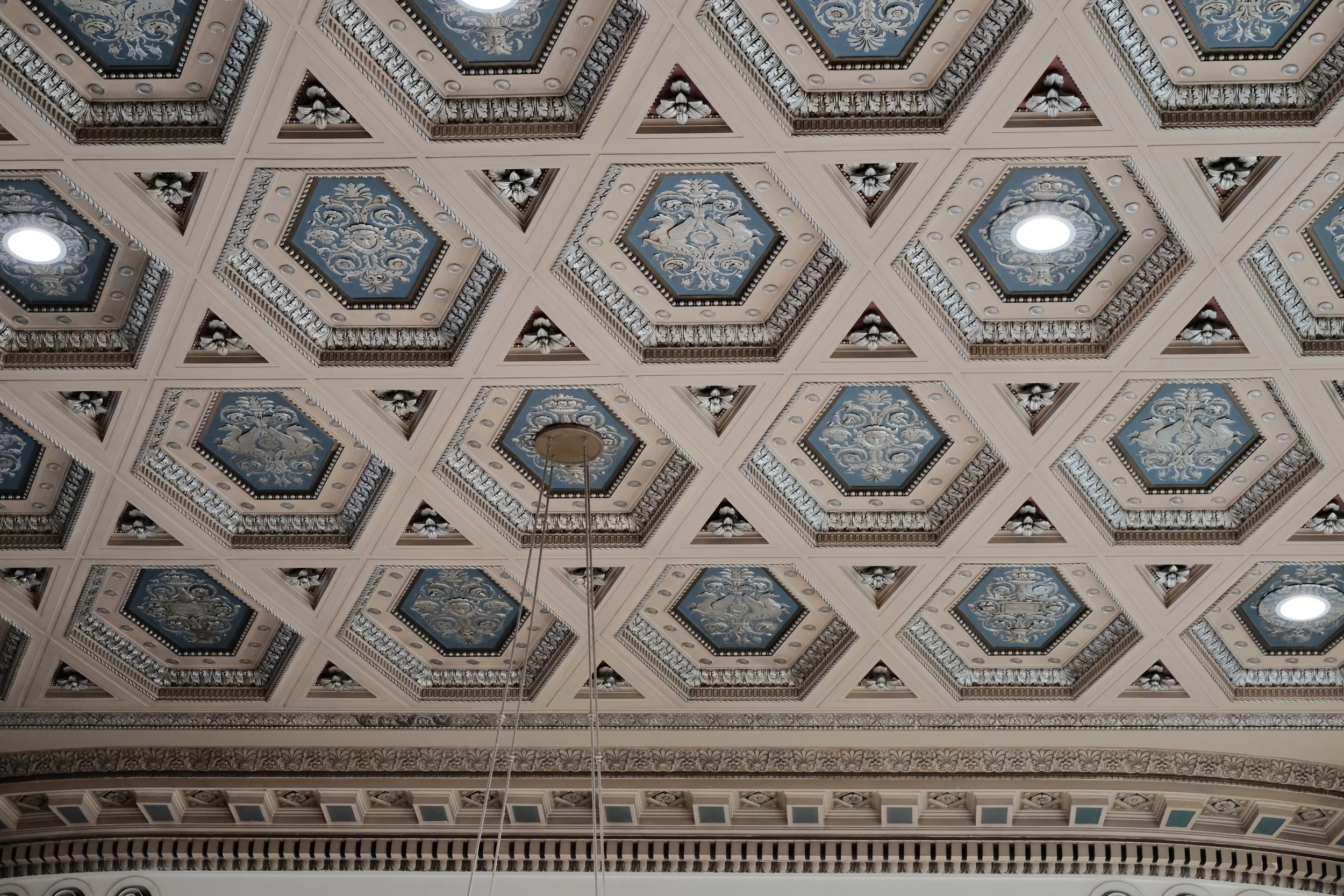 Decorative ornate ceiling with square panels, each featuring a painted floral design and surrounded by intricate moldings, with some recessed lighting fixtures.