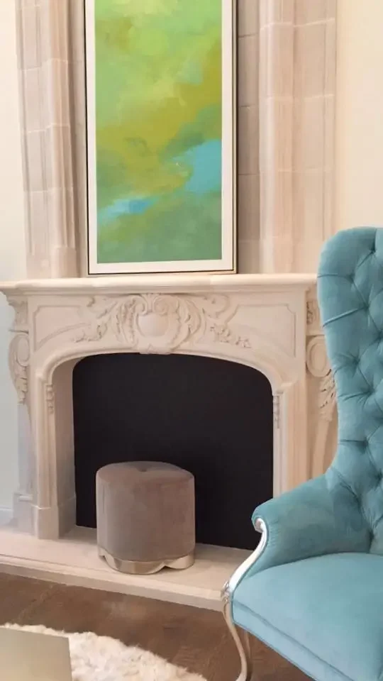 A white ornate fireplace with a black interior, topped with a colorful abstract painting with green, yellow, and blue hues. To the right, part of a light blue upholstered armchair is visible.