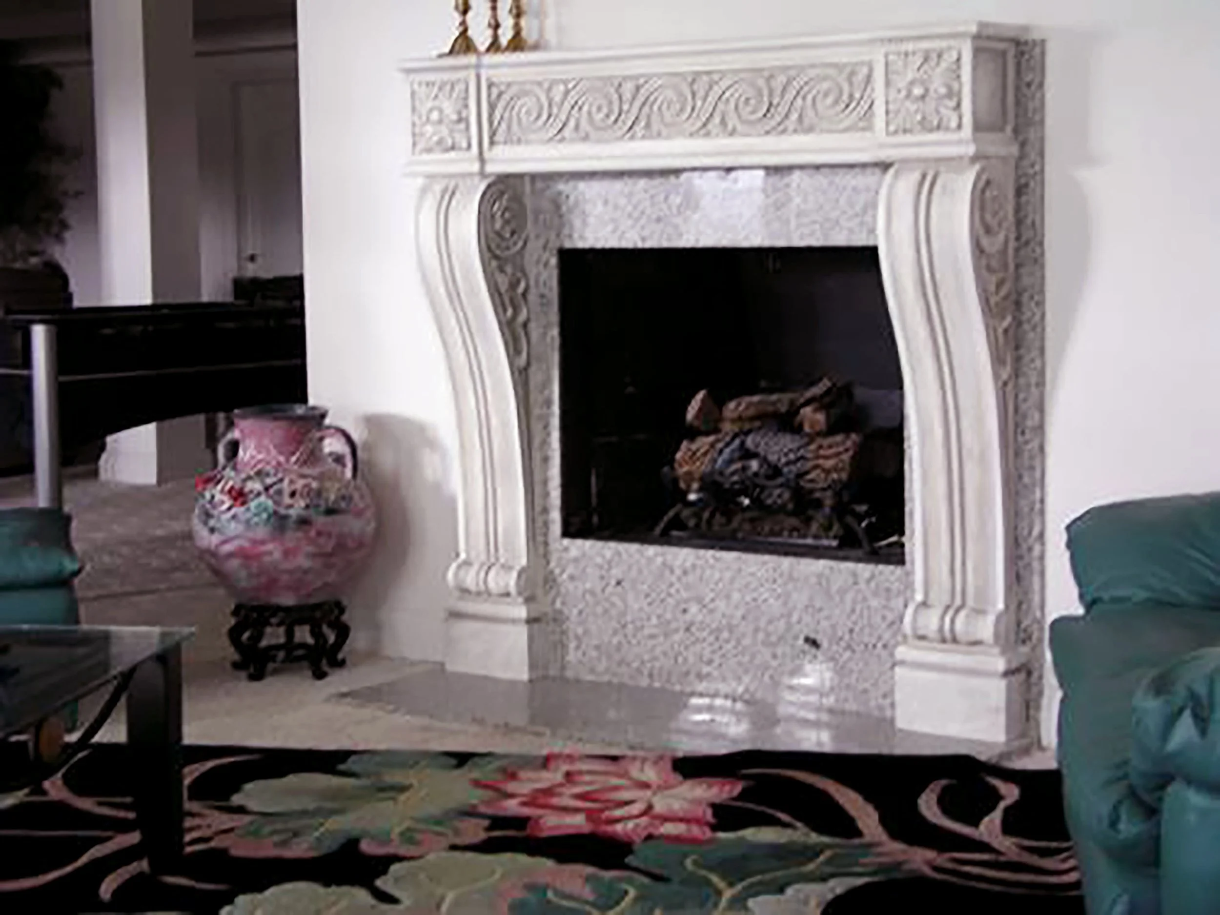 Living room with a marble fireplace, colorful vase, floral patterned rug, and partially visible green armchair.