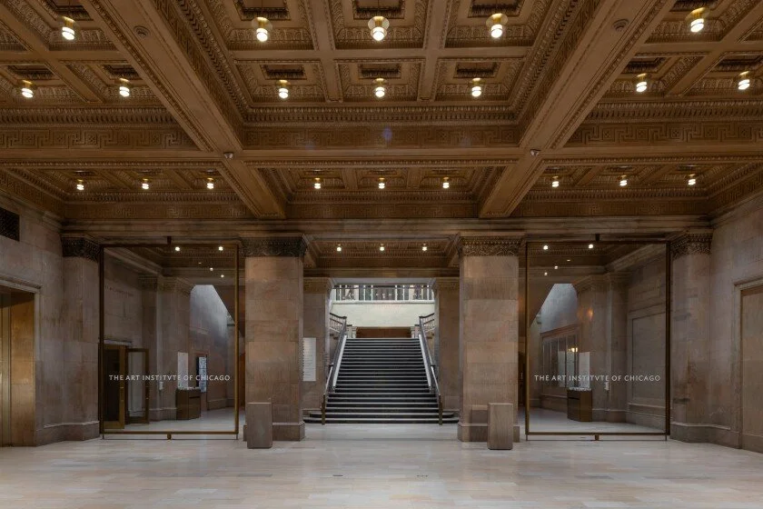 Interior of a gallery or lobby with grand staircase, ornate ceiling, and glass panels showing art displays from The Art Institute of Chicago.