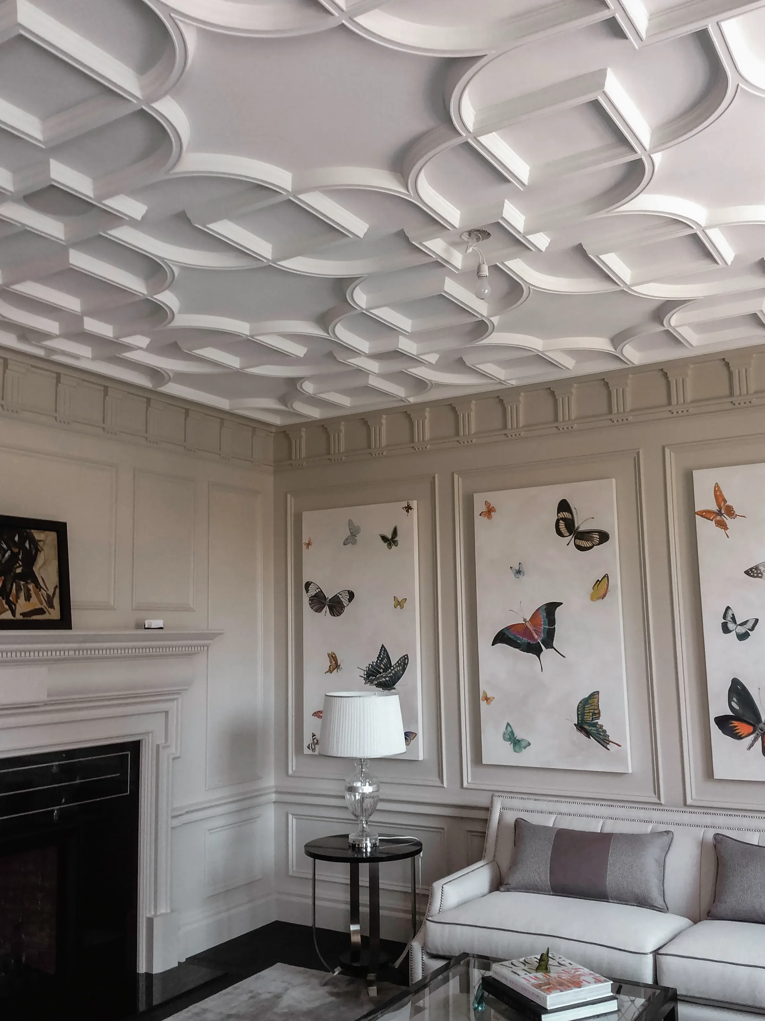 Living room with a white sofa, glass coffee table, and a side table with a lamp. Decorative wall panels with butterfly artwork and a marble fireplace. Coffered ceiling with geometric patterns.