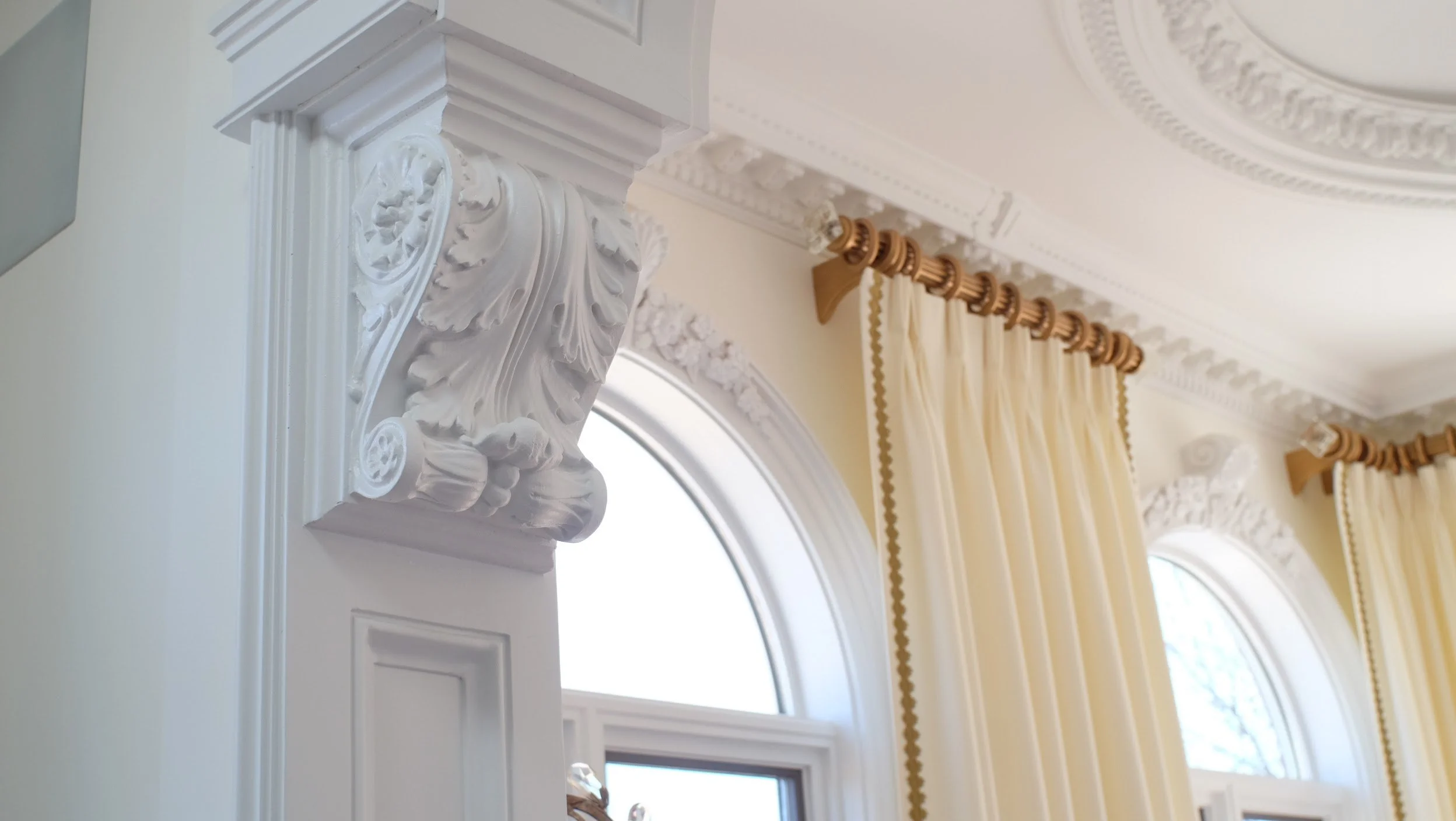 Close-up of ornate white crown molding and decorative plasterwork on ceiling and wall in a room with large arched windows and beige curtains with gold trim.