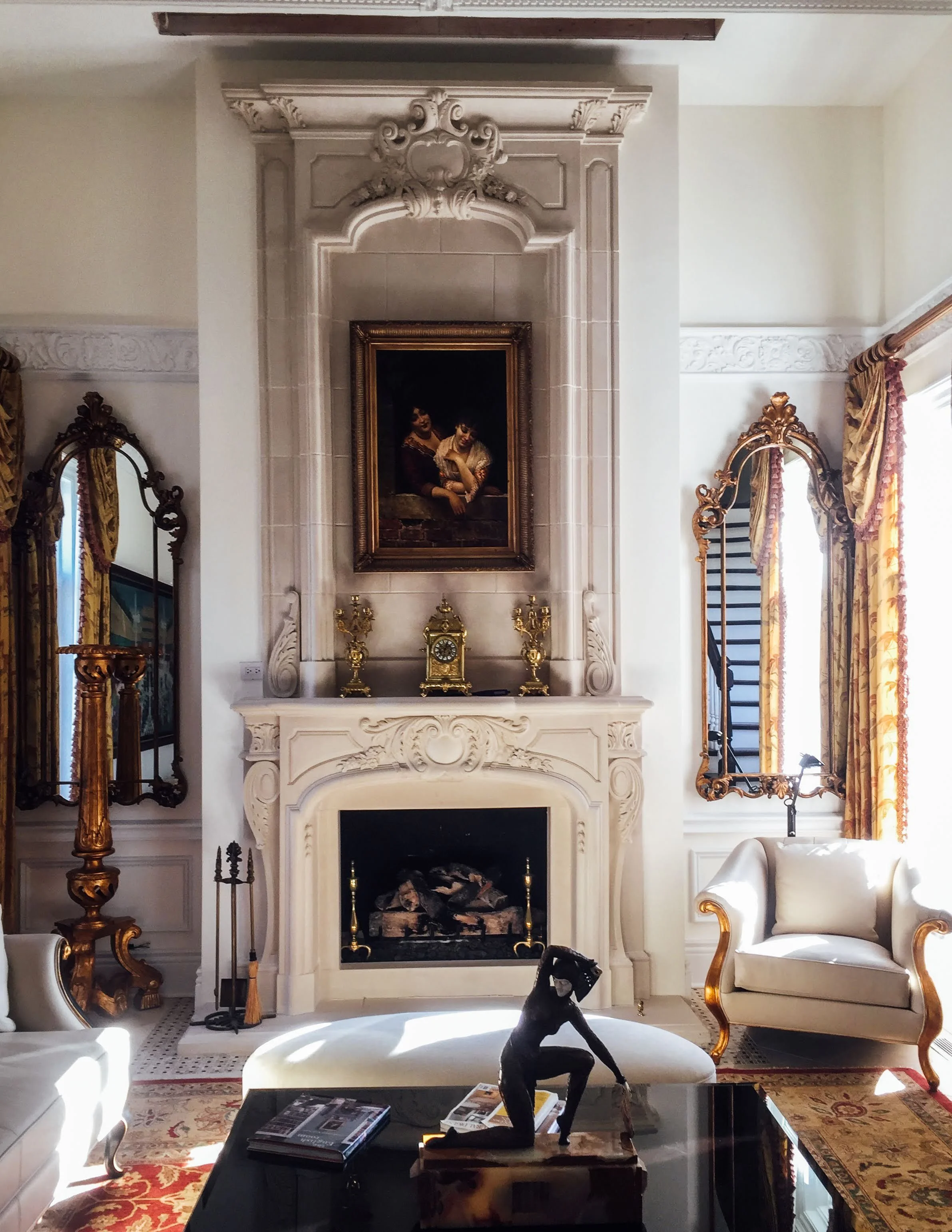 Elegant living room with a white ornate fireplace, a painting of two women above it, gold decorative objects on the mantel, and a black sculpture of a woman on the coffee table. There are vintage mirrors with gold frames, a white armchair, a dark woo