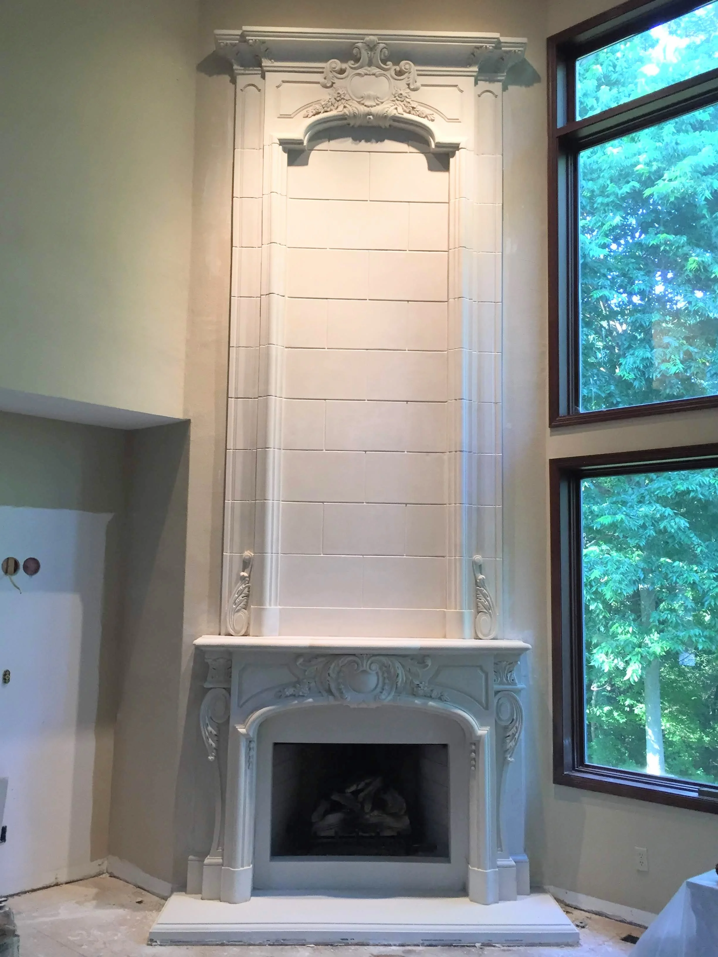 White ornate fireplace with decorative molding and carvings, situated between two large windows showing green trees outside.